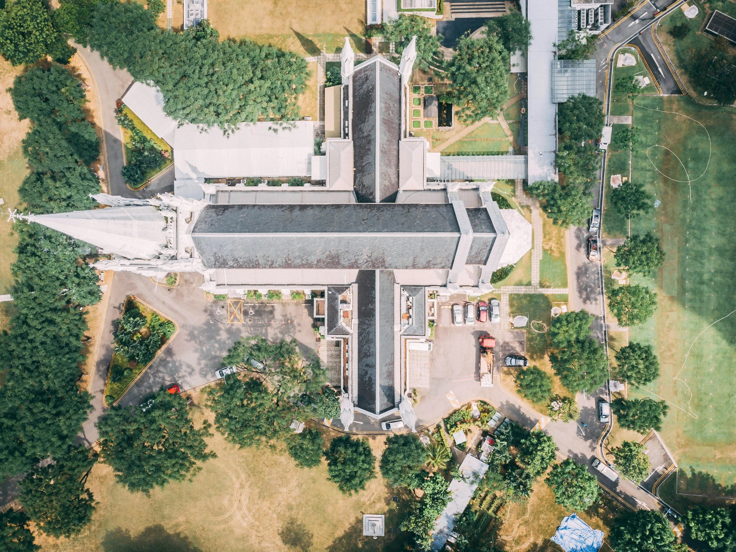 Vue aérienne d'une église ancienne entourée d'arbres, de jardins et d'espaces verts, avec des parkings et un terrain de sport à proximité.