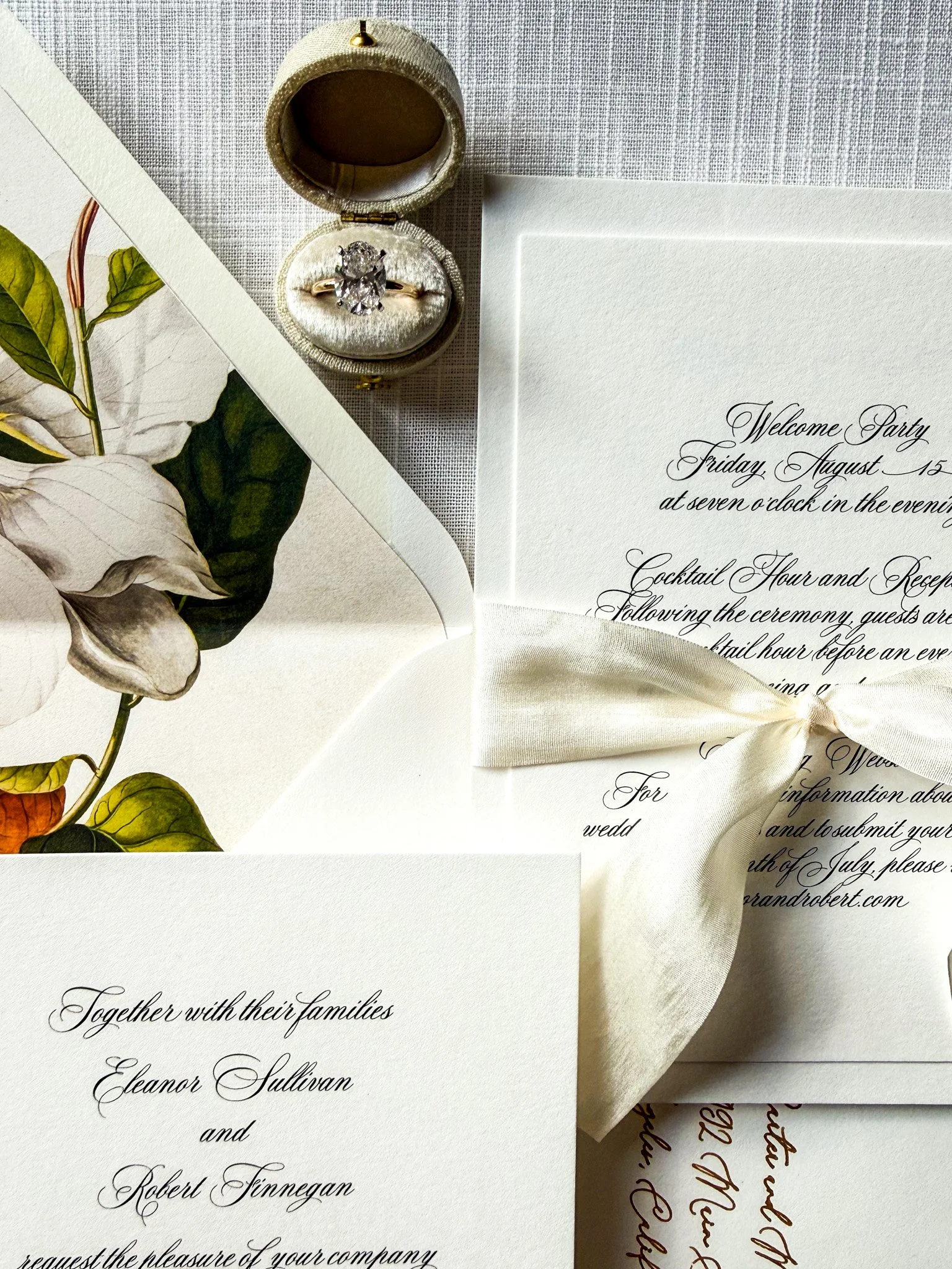 A wedding invitation suite with calligraphy, a white satin ribbon, a ring box with a diamond ring, a botanical illustration, and a white textured background.