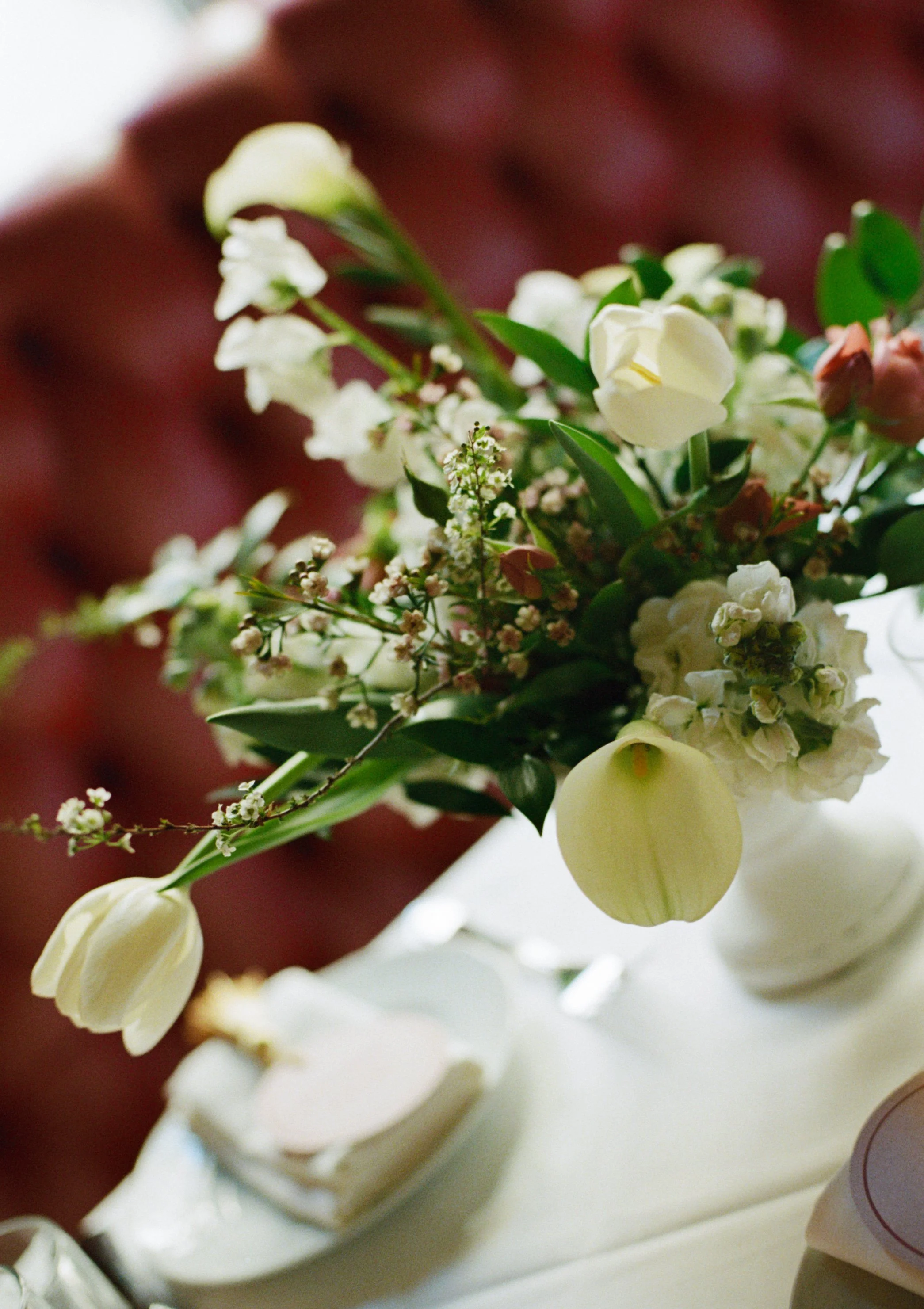 Close-up of a bouquet of white and light pink flowers with green leaves, placed in a white vase on a white surface.