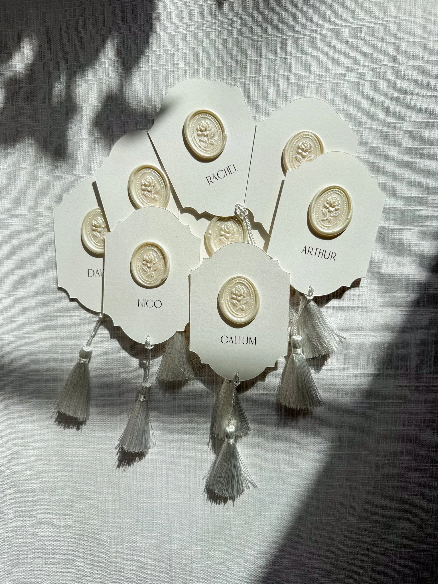 White place cards, each having an oval wax seal embossed with a rose design and names written in black, such as RACHEL, ARTHUR, NICO, CALLUM, DAPHNE, and others. The bottom of each tag has white tassels.