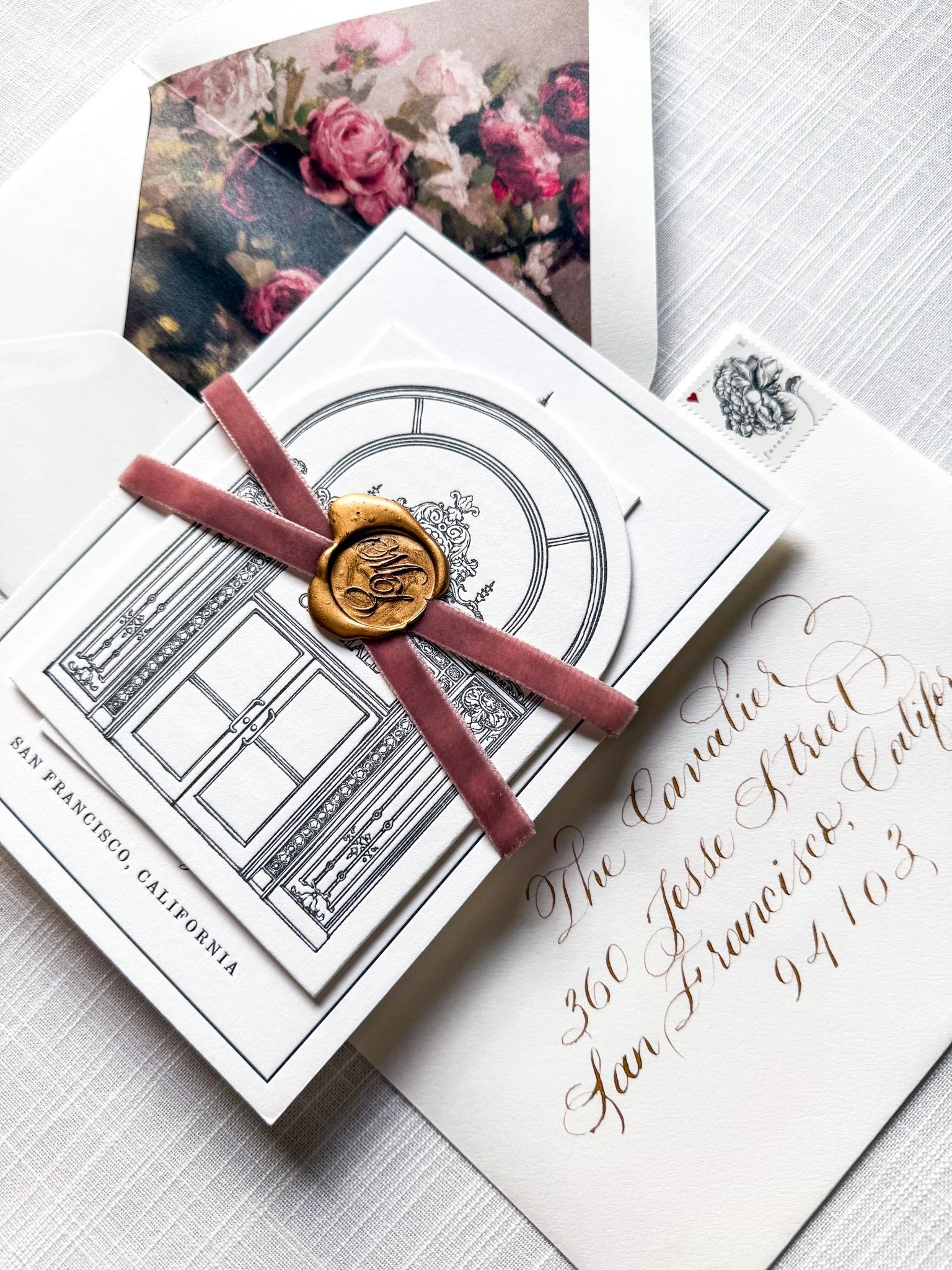 A wedding invitation suite with a gold wax seal featuring the letters 'M' and 'Z', a pink velvet ribbon, and a black and white illustration of an arched doorway, partially covering a floral photo and handwritten letter in elegant cursive.
