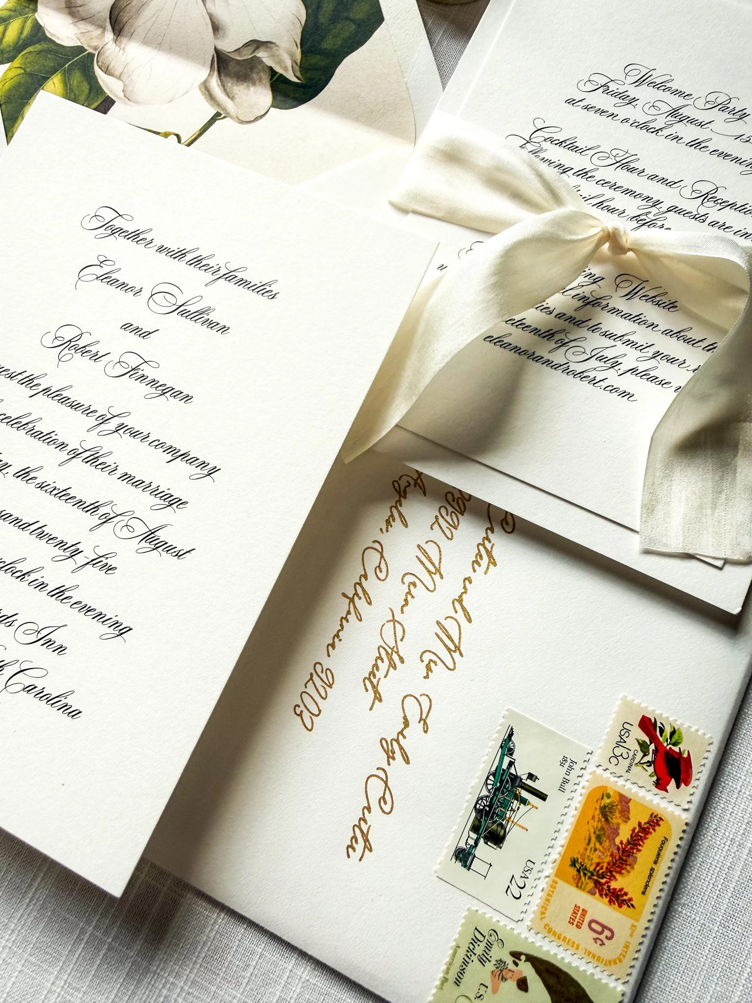 Wedding invitation with black cursive text, floral illustration, cream-colored ribbon, and envelope with vintage postage stamps, on light-colored fabric background.