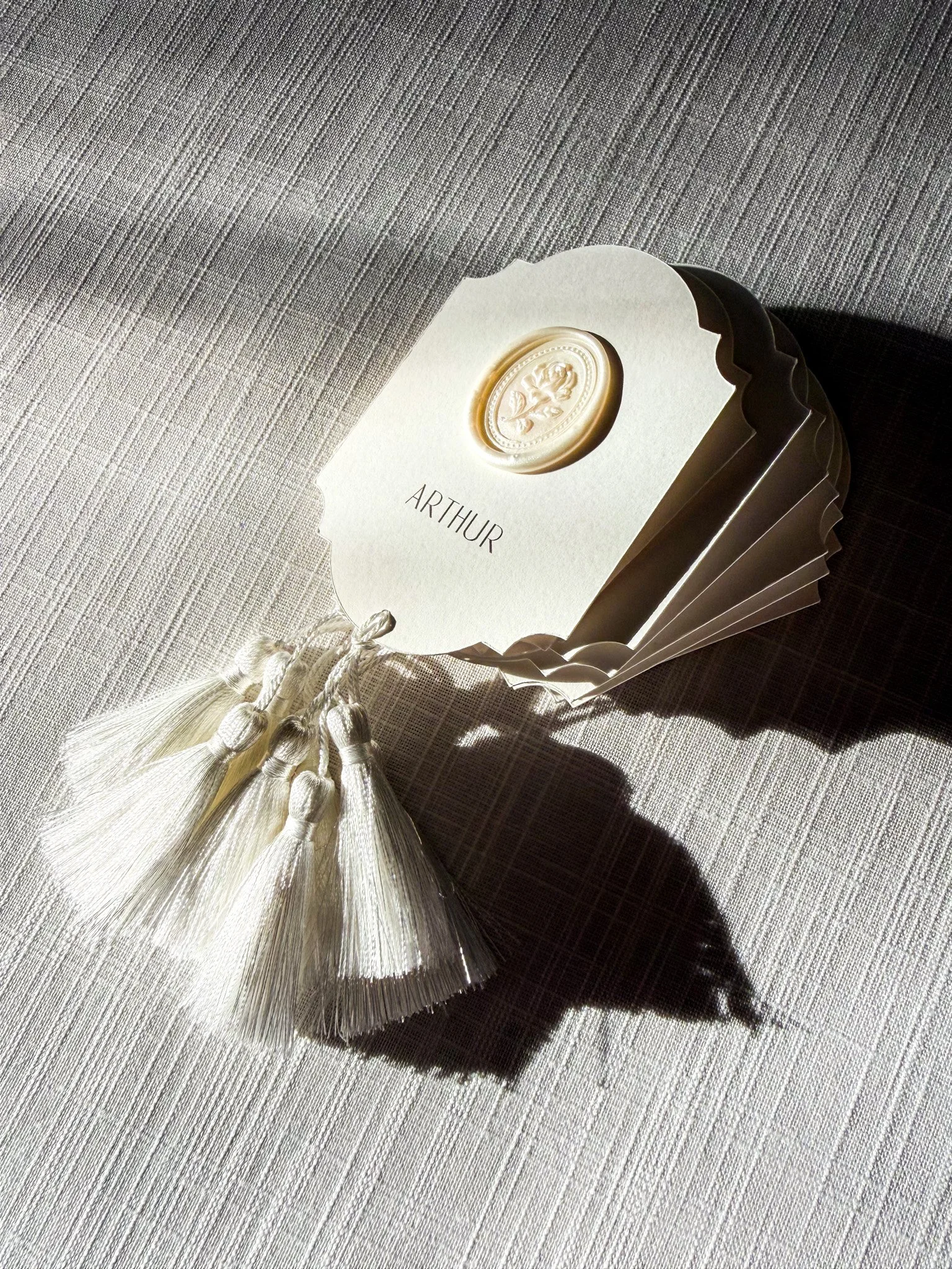 A set of cream-colored invitation cards secured with a decorative tassel and seal, featuring the name 'Arthur' in elegant black font, placed on a textured white surface.
