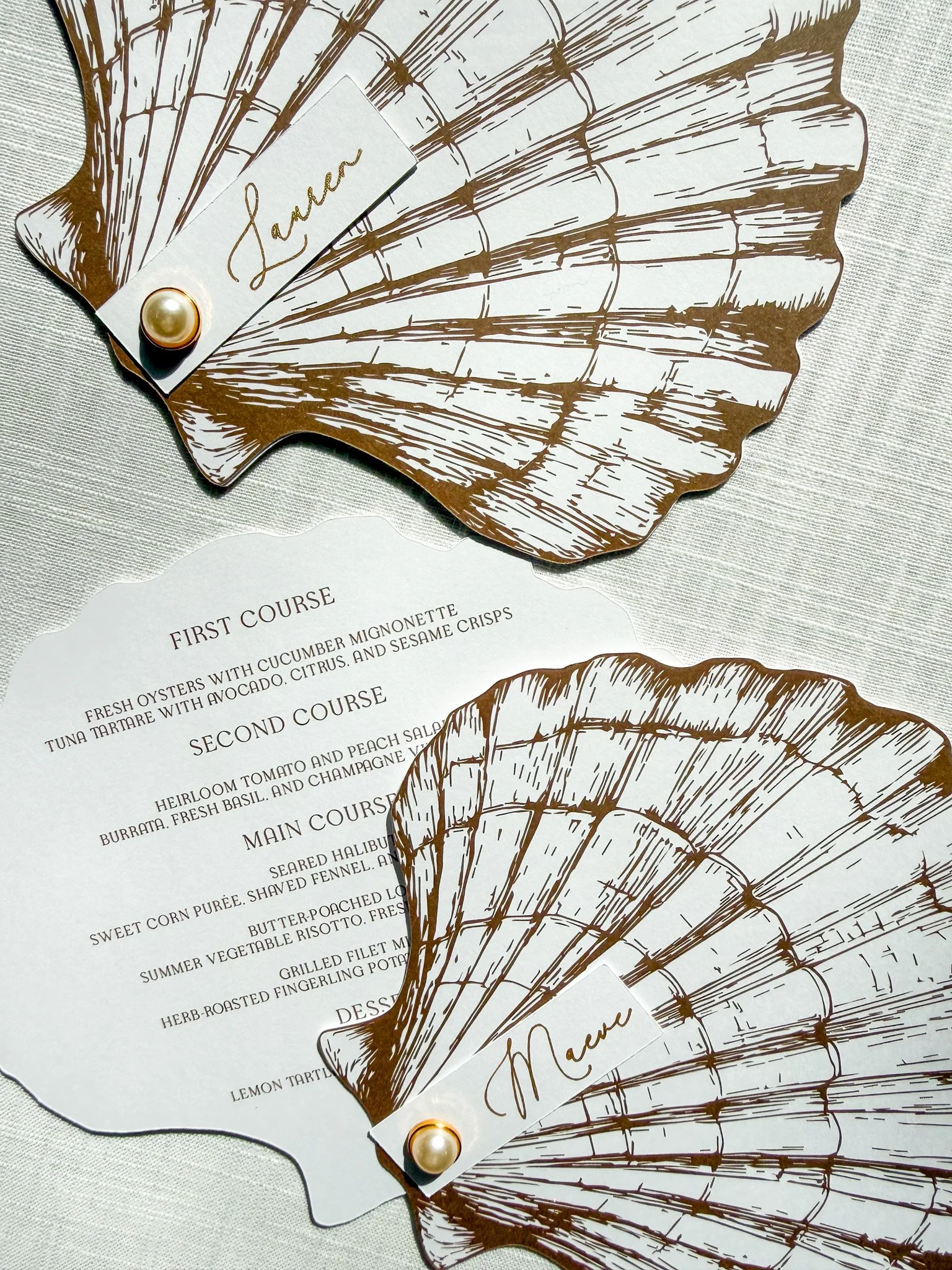 A decorative paper fan with a white background and brown line drawing of a fan shape. Attached to the fan is a small white card with the name 'Sarah' written in gold script, secured with a gold pearl pin. A menu with dishes and courses is partially v