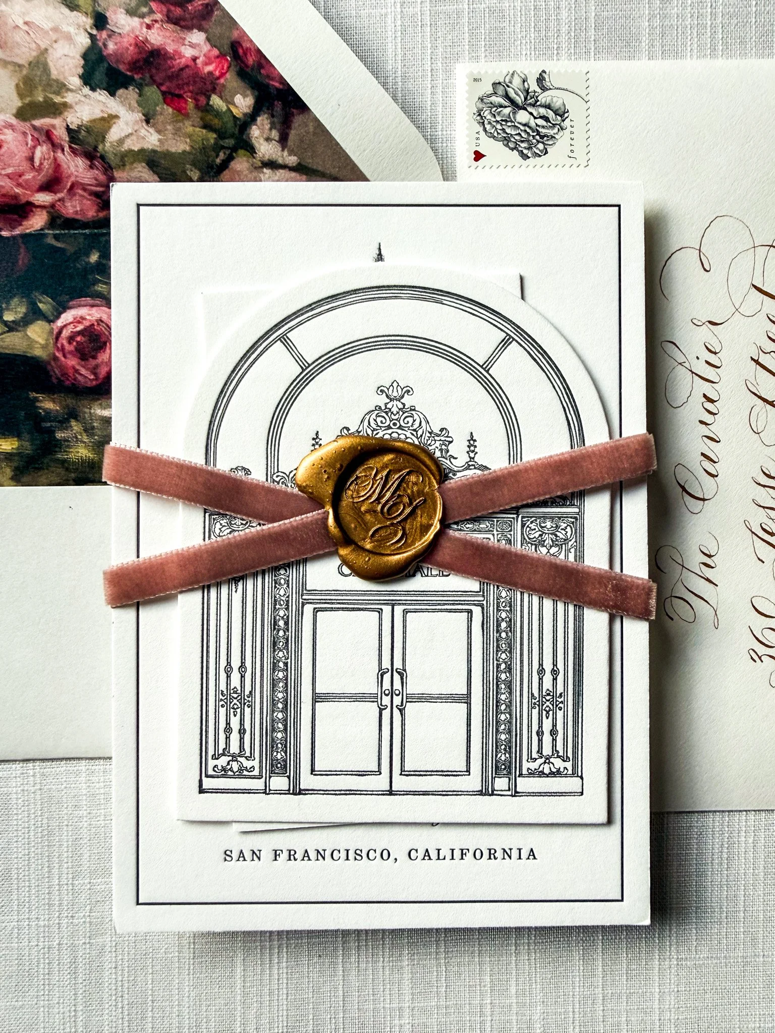 A wedding invitation card with a pink satin ribbon and a gold wax seal. The card features an engraved door illustration at the top and the words "San Francisco, California" at the bottom. Part of a floral envelope with pink roses and a postage stamp 
