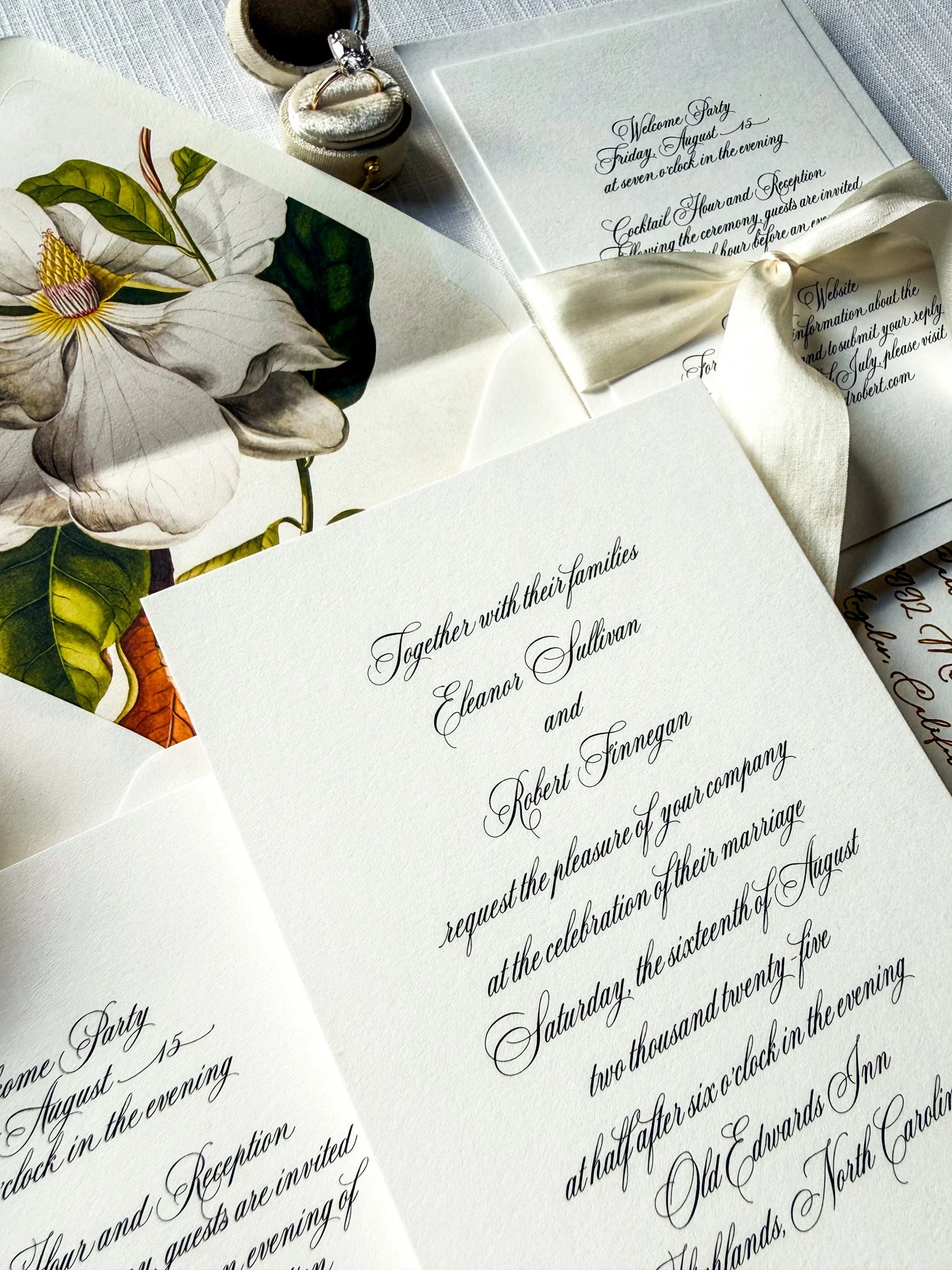 Close-up of wedding invitations with elegant black cursive font, floral artwork, a satin ribbon, and wedding rings, laid on a white textured fabric.