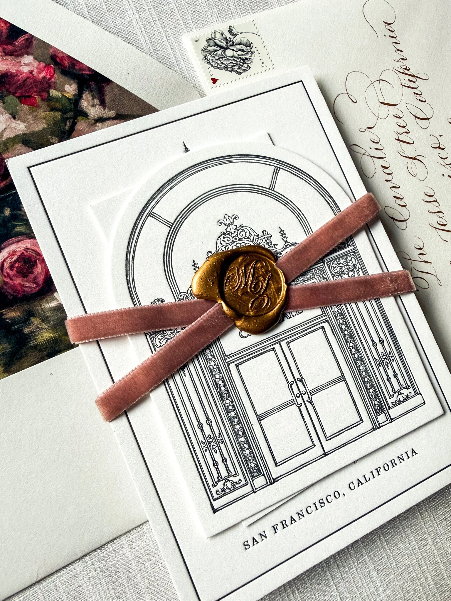 A wedding invitation with an illustration of a door, a pink velvet ribbon, a gold wax seal with a monogram, a piece of paper with elegant handwritten calligraphy, and a small postage stamp featuring a rose illustration.