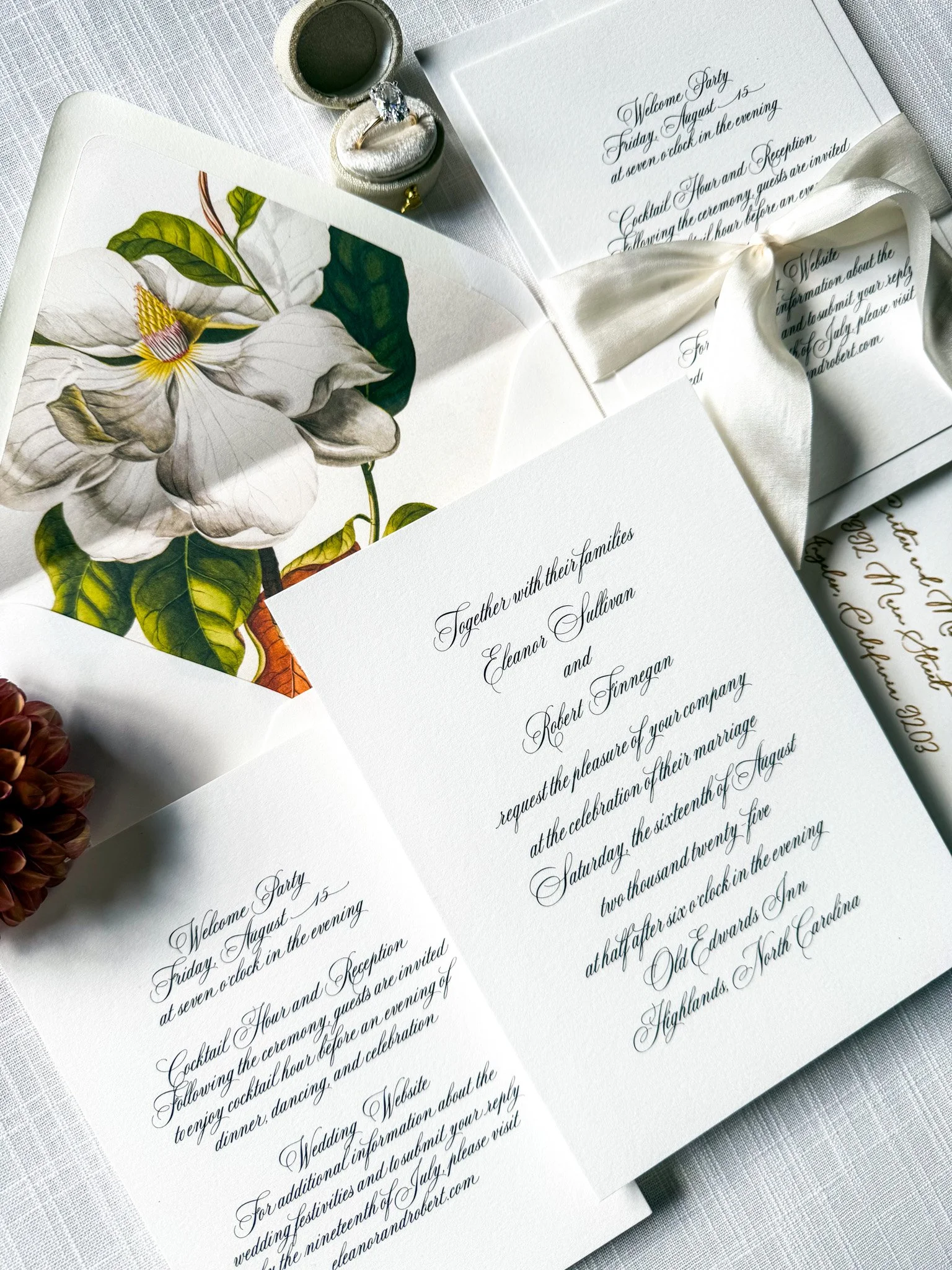 Wedding invitation cards with elegant calligraphy text, a white satin ribbon, and a small ring box containing a diamond ring. The cards feature floral artwork and event details.