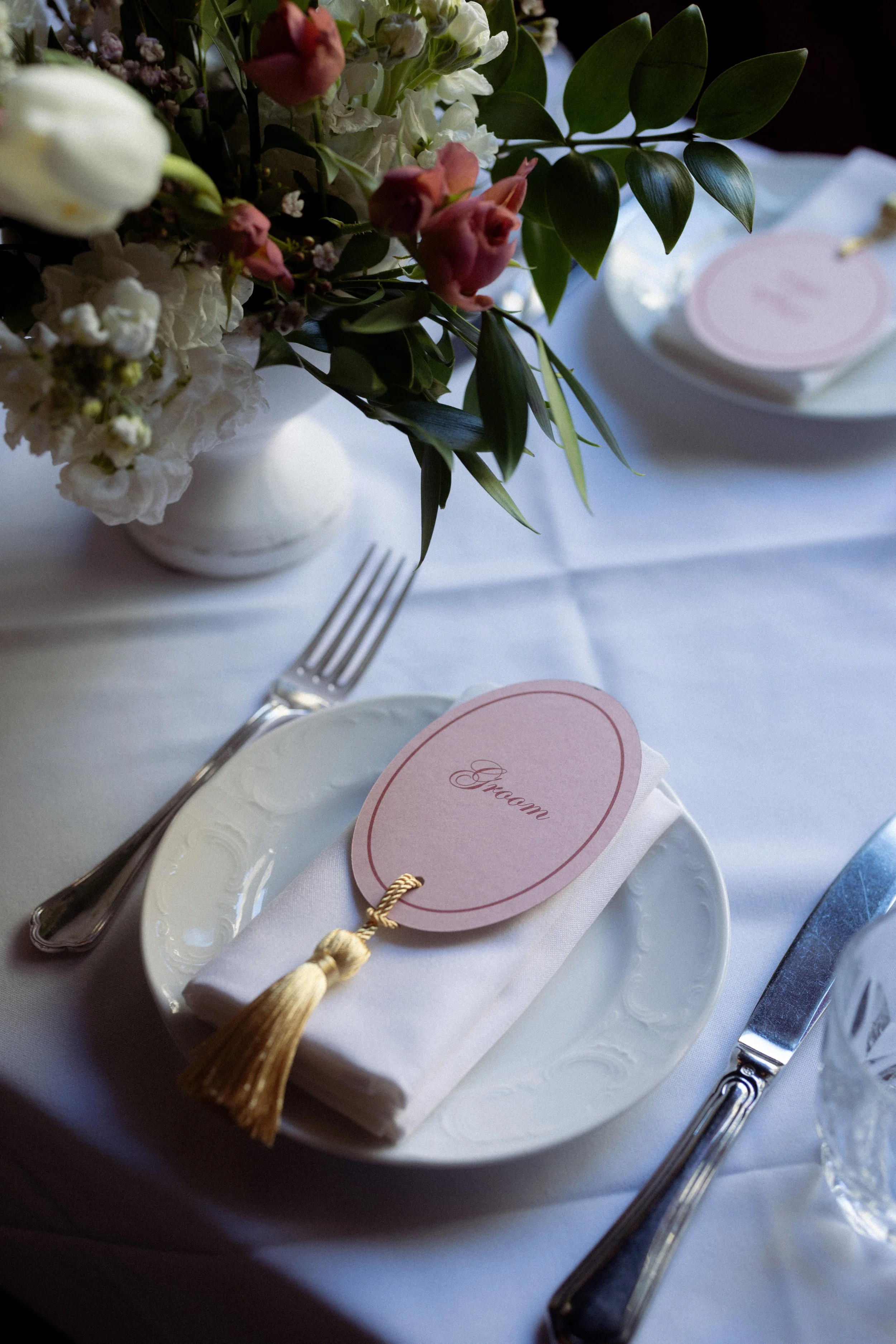 A place setting at a formal event featuring a white plate, a white napkin with a pink 'Groom' tag attached and a gold tassel, a fork, and a knife. In the background, there is a floral arrangement with white and pink flowers and green leaves.