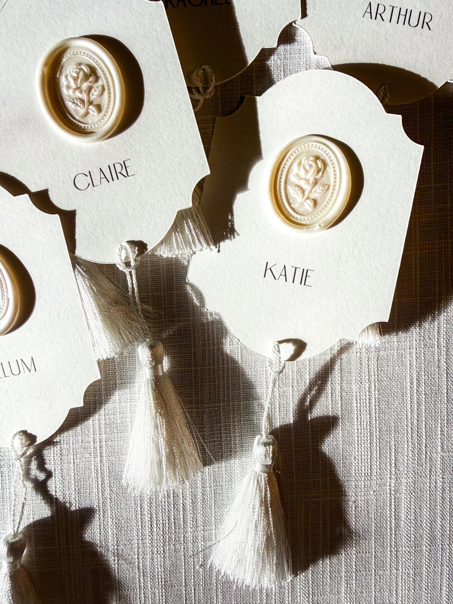 Close-up of personalized wax seals on blank cards labeled 'Claire', 'Katie', and 'Arthur'. The cards are placed on a textured fabric surface, with shadows and light creating a warm, elegant atmosphere.