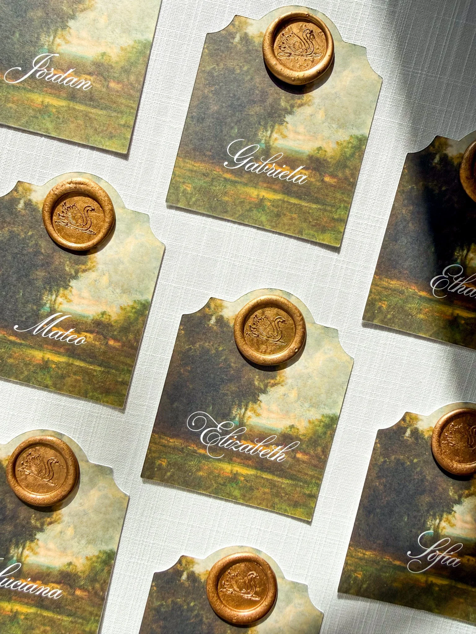 Place cards with names 'Gabriele', 'Matteo', and 'Elizabeth' are arranged on a table. Each card has a landscape painting background and a gold seal with a swan emblem.