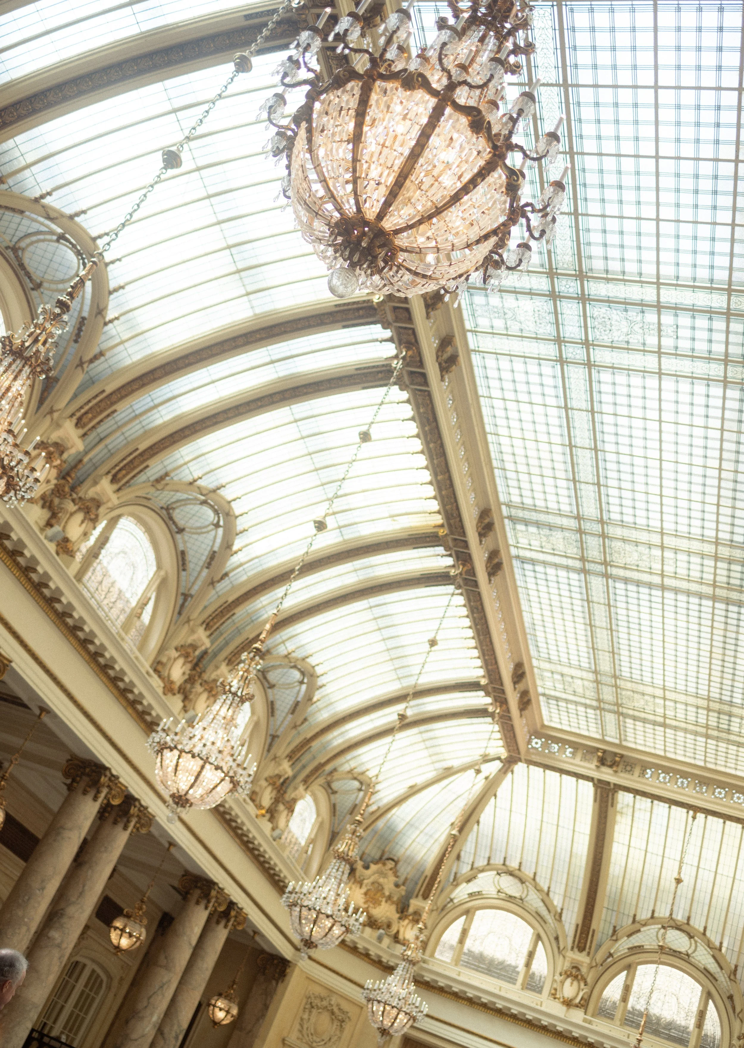 Interior view of a grand hall with large crystal chandeliers hanging from a high glass ceiling with ornate gold and white decor.