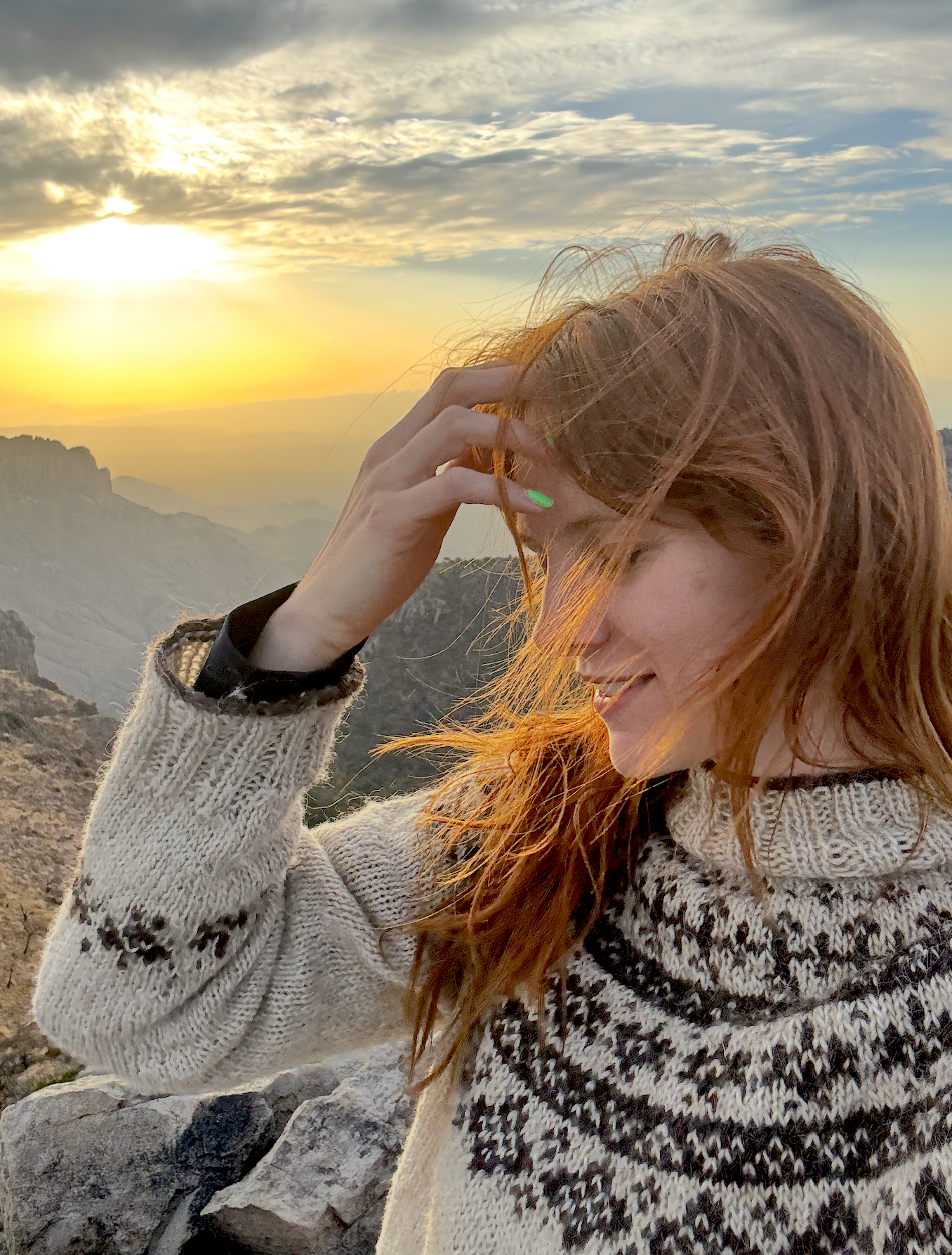 A woman with red hair and a pull-over sweater smiling and touching her hair, with a mountain landscape and sunset in the background.