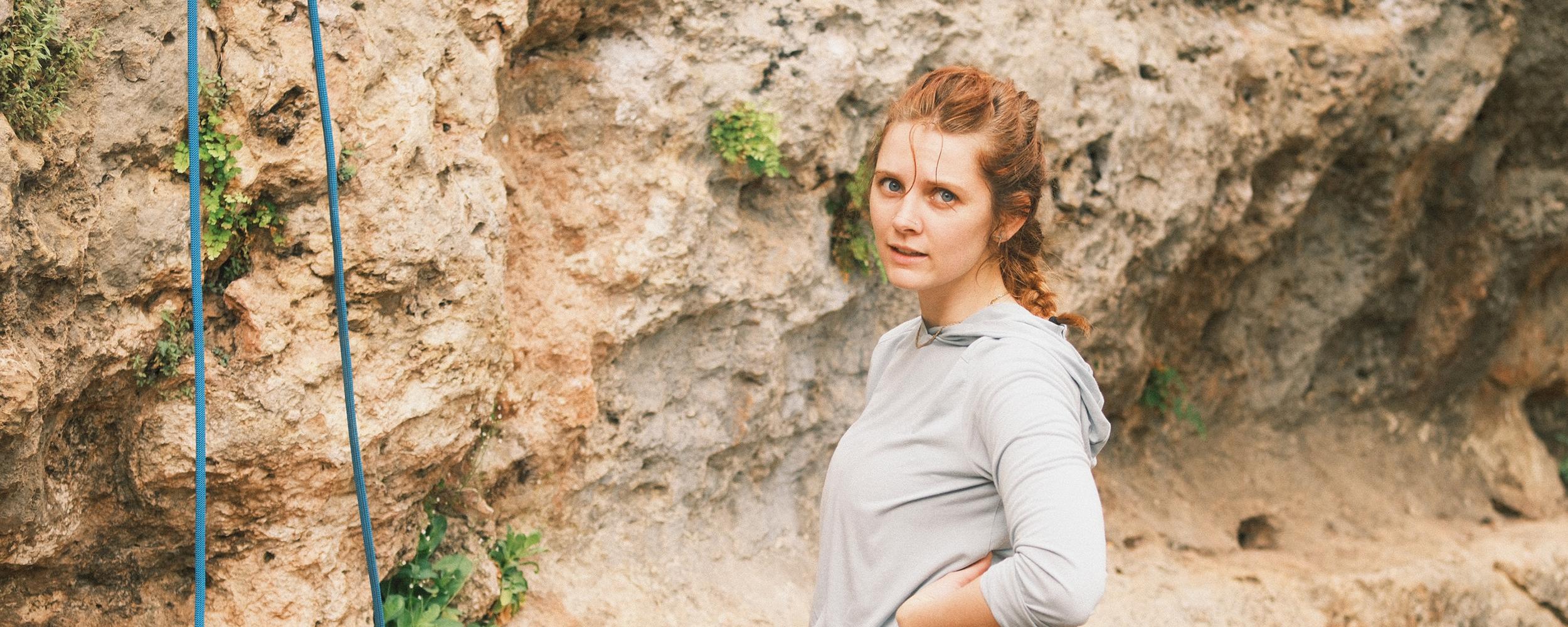 Young woman with red hair in a braid, wearing a light gray hoodie, standing outdoors in front of a rocky cliff, looking at the camera with a curious expression.