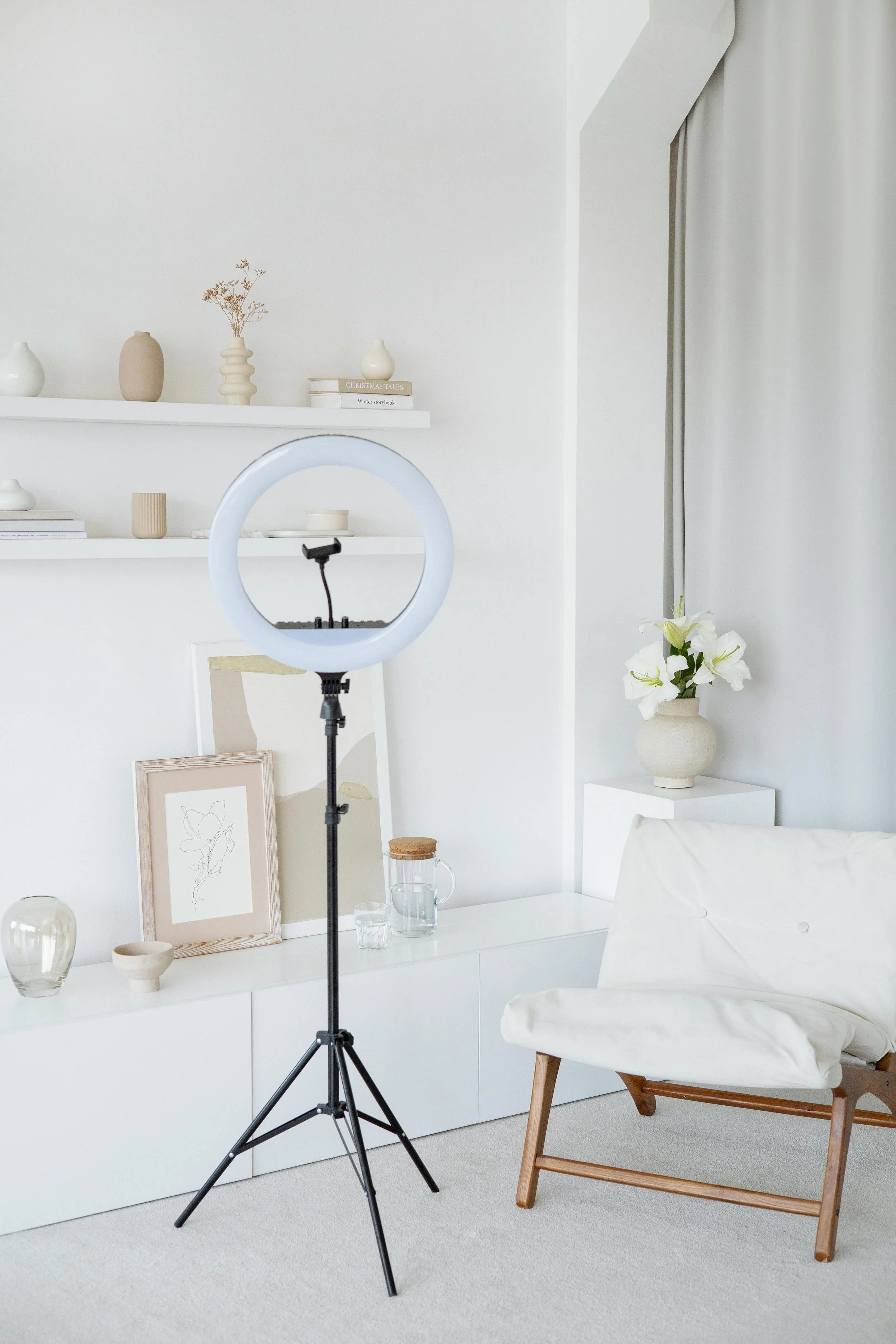A white bedroom with a large ring light on a black stand in front of a chair and a bed with hanging white curtains, decorated with beige vases, books, and framed artwork.