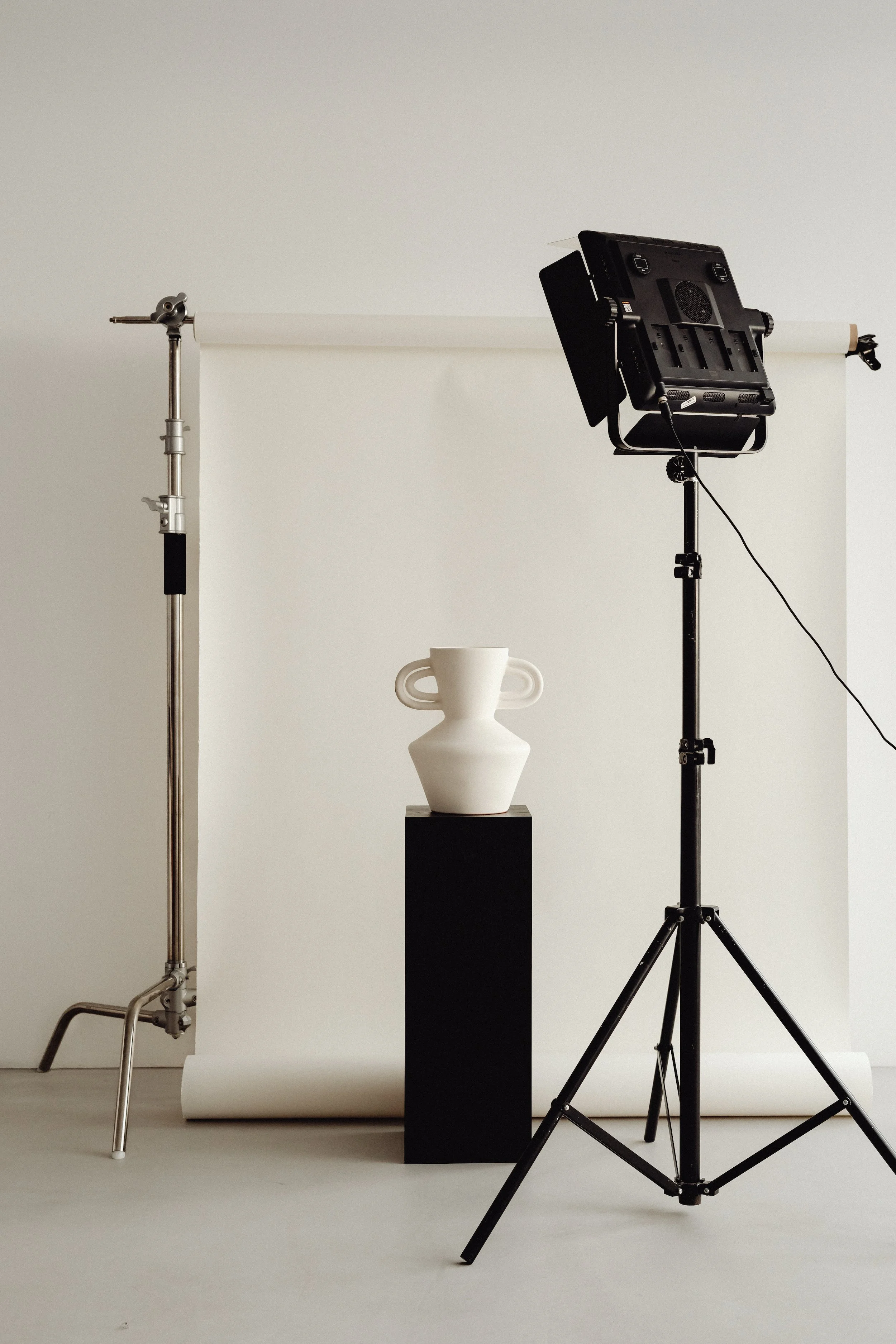 Photography studio setup with a white backdrop, a white ceramic vase with handles on a black pedestal, a large softbox light, a reflector, and lighting stands on a smooth white floor.