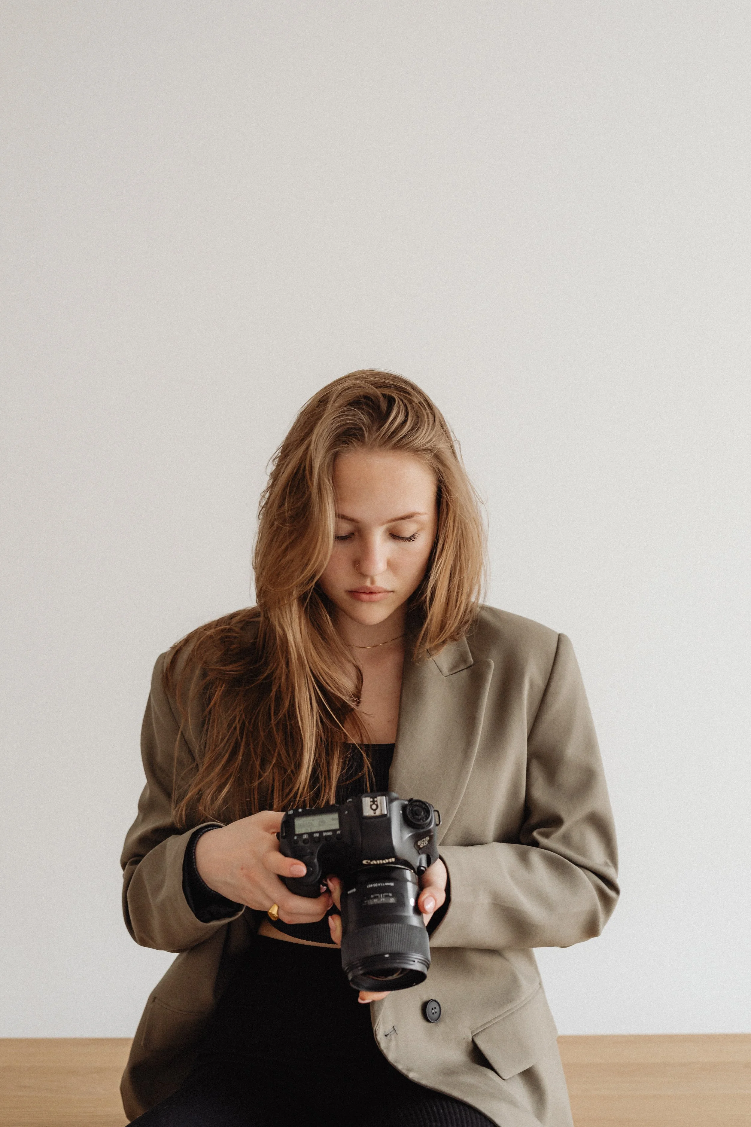 A young woman with long, wavy, light brown hair holding a professional DSLR camera with a large lens, looking down at it, dressed in a beige blazer and black top, seated in front of a plain white wall.