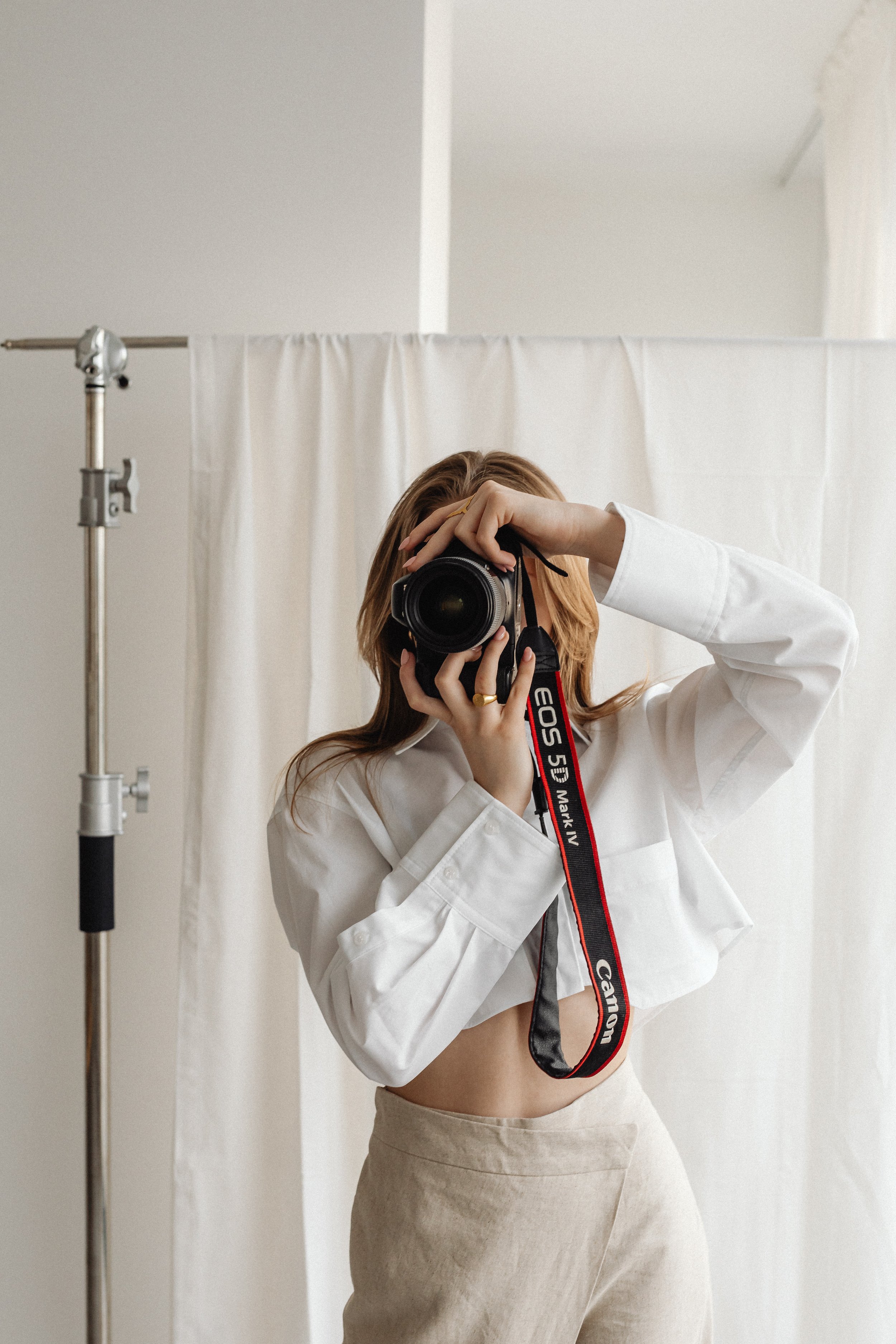 Woman taking a selfie with a camera in a photography studio, wearing a white shirt and beige pants, with a white backdrop and studio equipment visible.