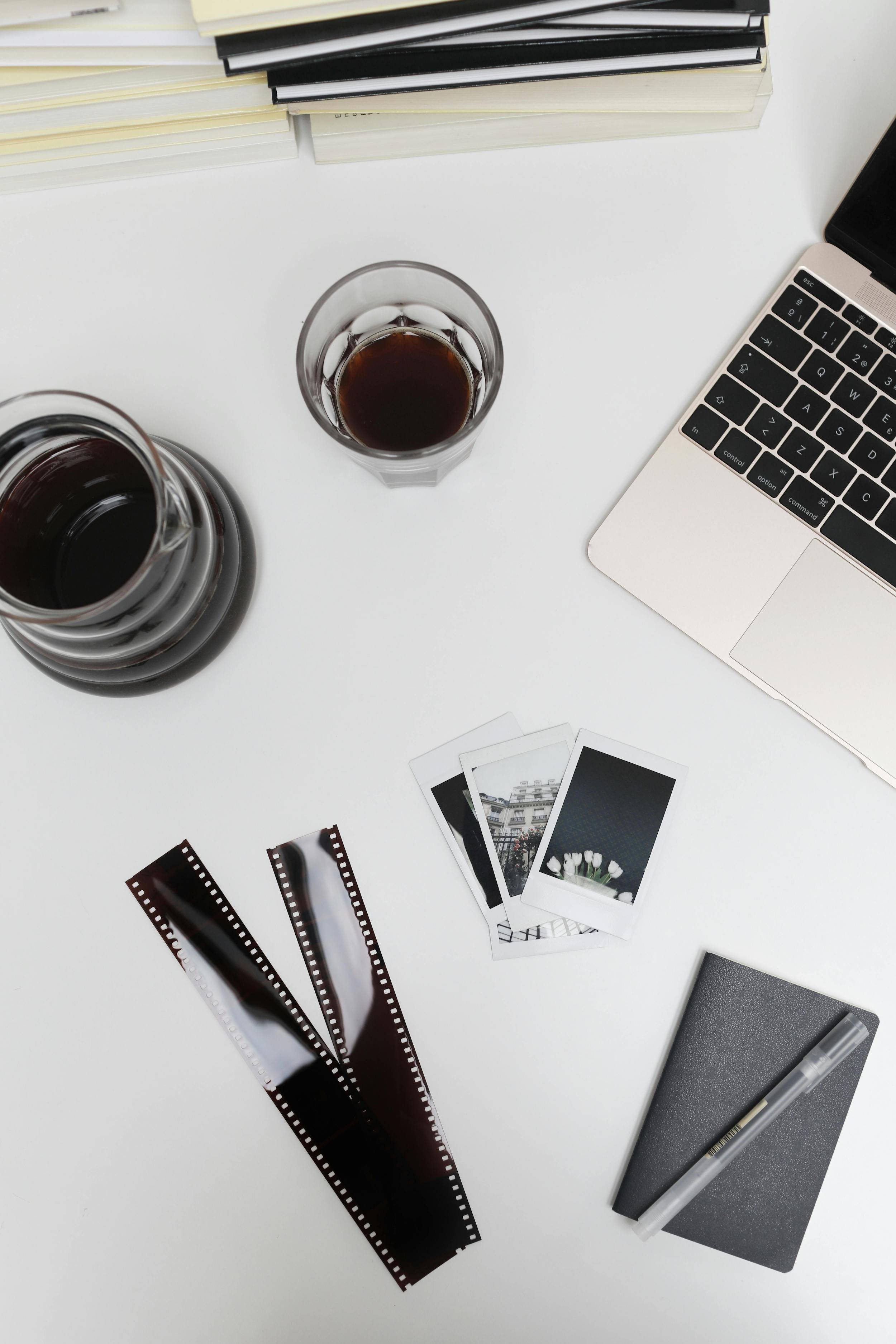 A white desk with three glasses of dark-colored soda, a laptop, books, a small notebook, a pen, photographs, and film strips.