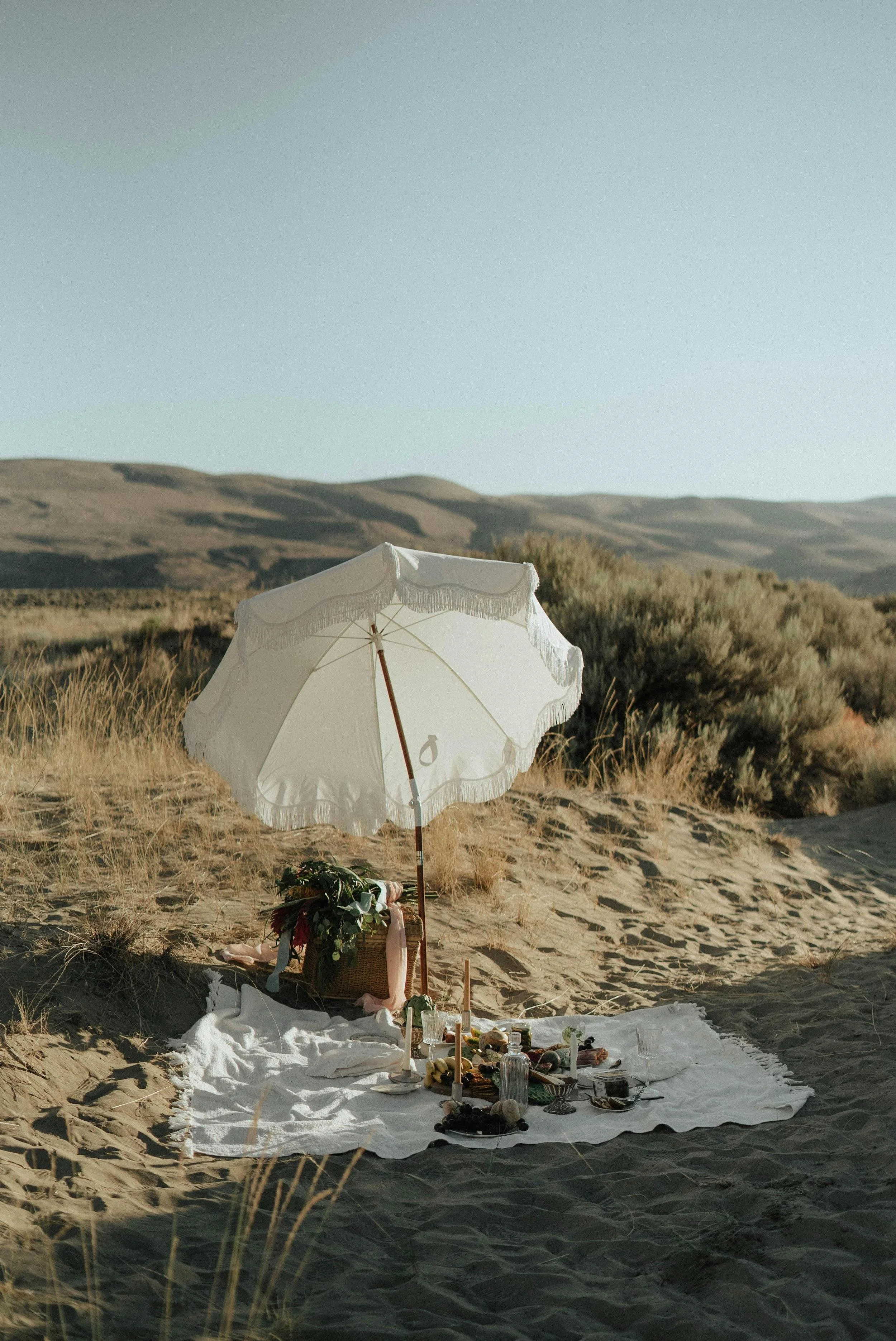 A picnic setup on the sand in a desert landscape with rolling hills in the background. There is a large white parasol, a white blanket with food, drinks, and flowers arranged on it, and a basket placed nearby.