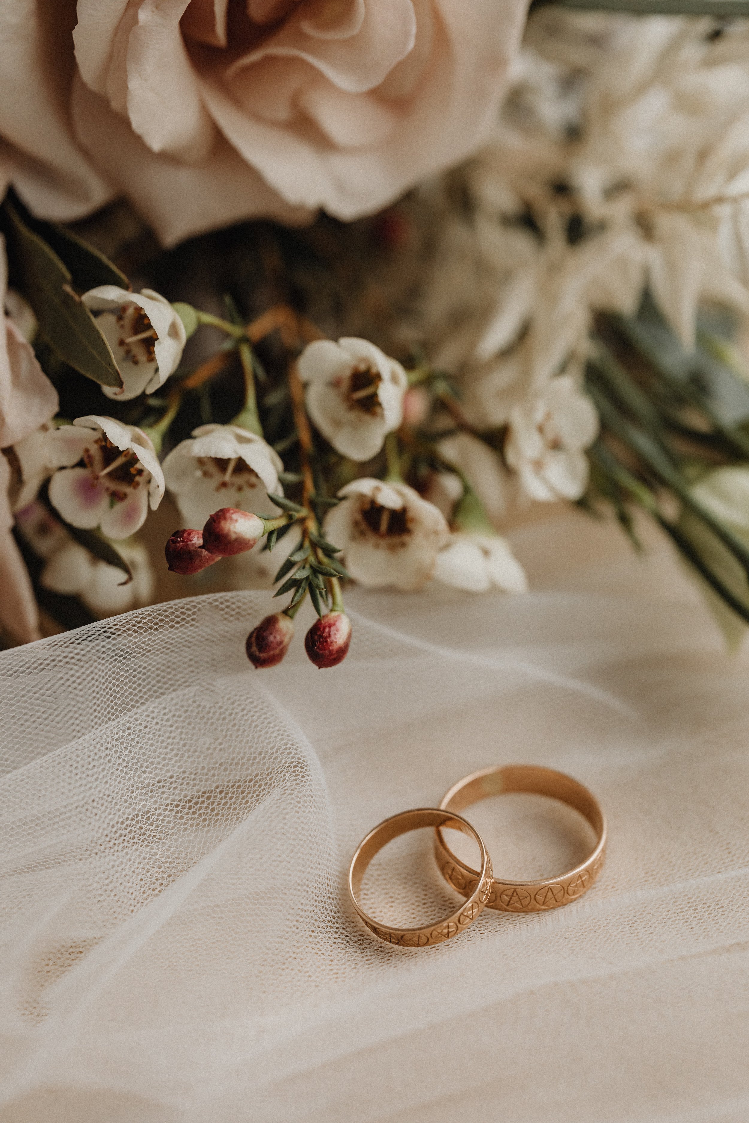 Close-up of two gold rings with engraved patterns placed on a white fabric, with a bouquet of white and pink flowers, including roses and small berry-like buds, in the background.