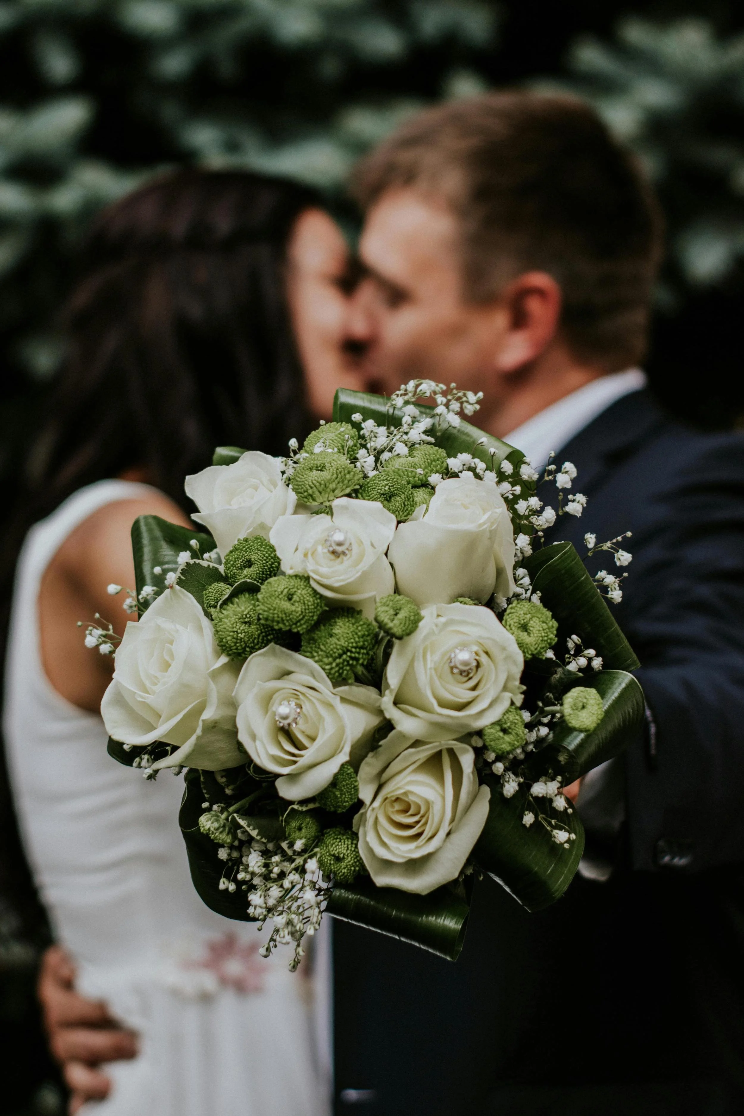 A bride holding a bouquet of white roses, baby's breath, and green flowers, with a groom in a suit in background at a wedding.