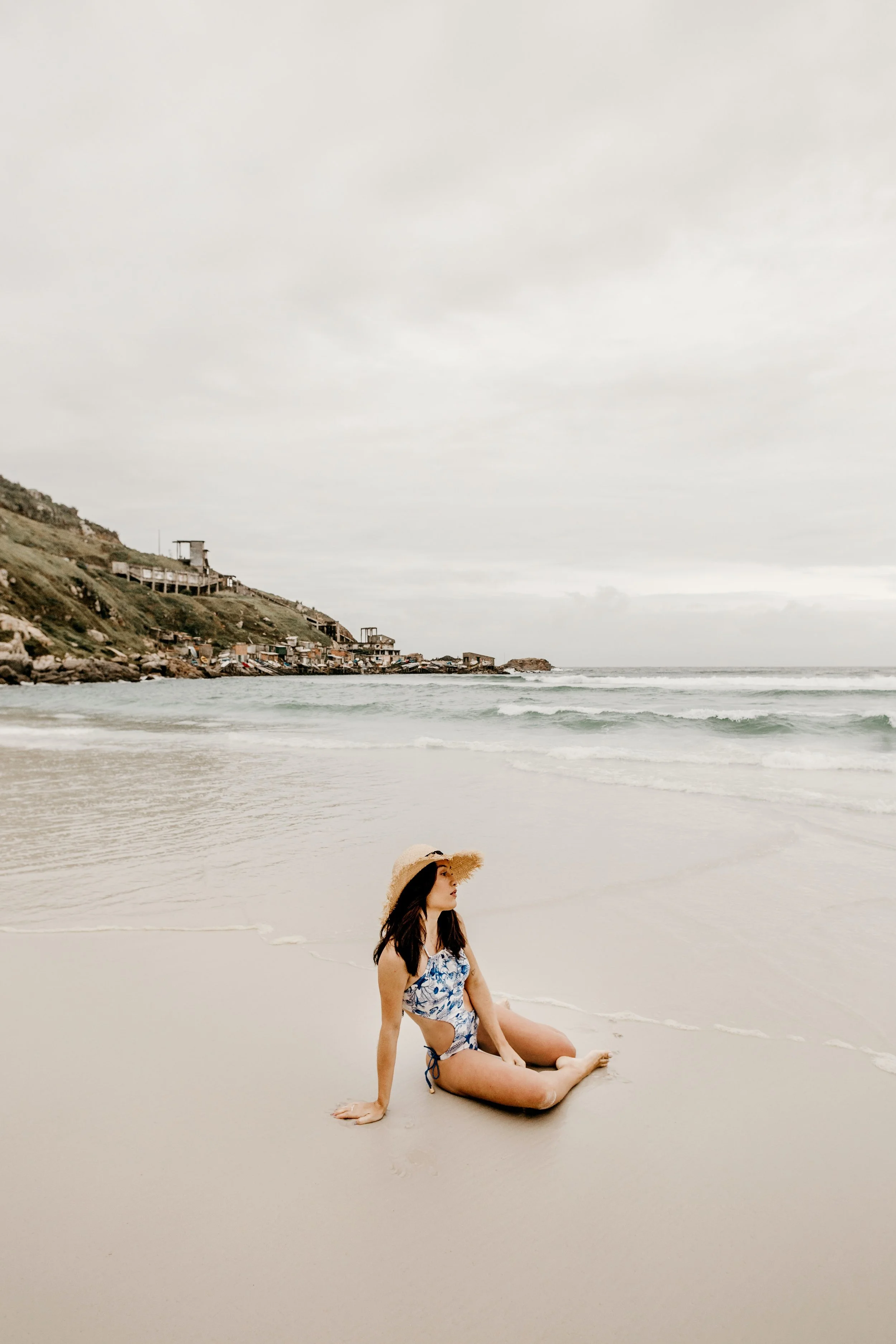 A woman with dark hair wearing a blue and white swimsuit and a wide-brimmed straw hat sitting on a sandy beach near the shore, with a coastal village with houses and a hillside in the background under a cloudy sky.