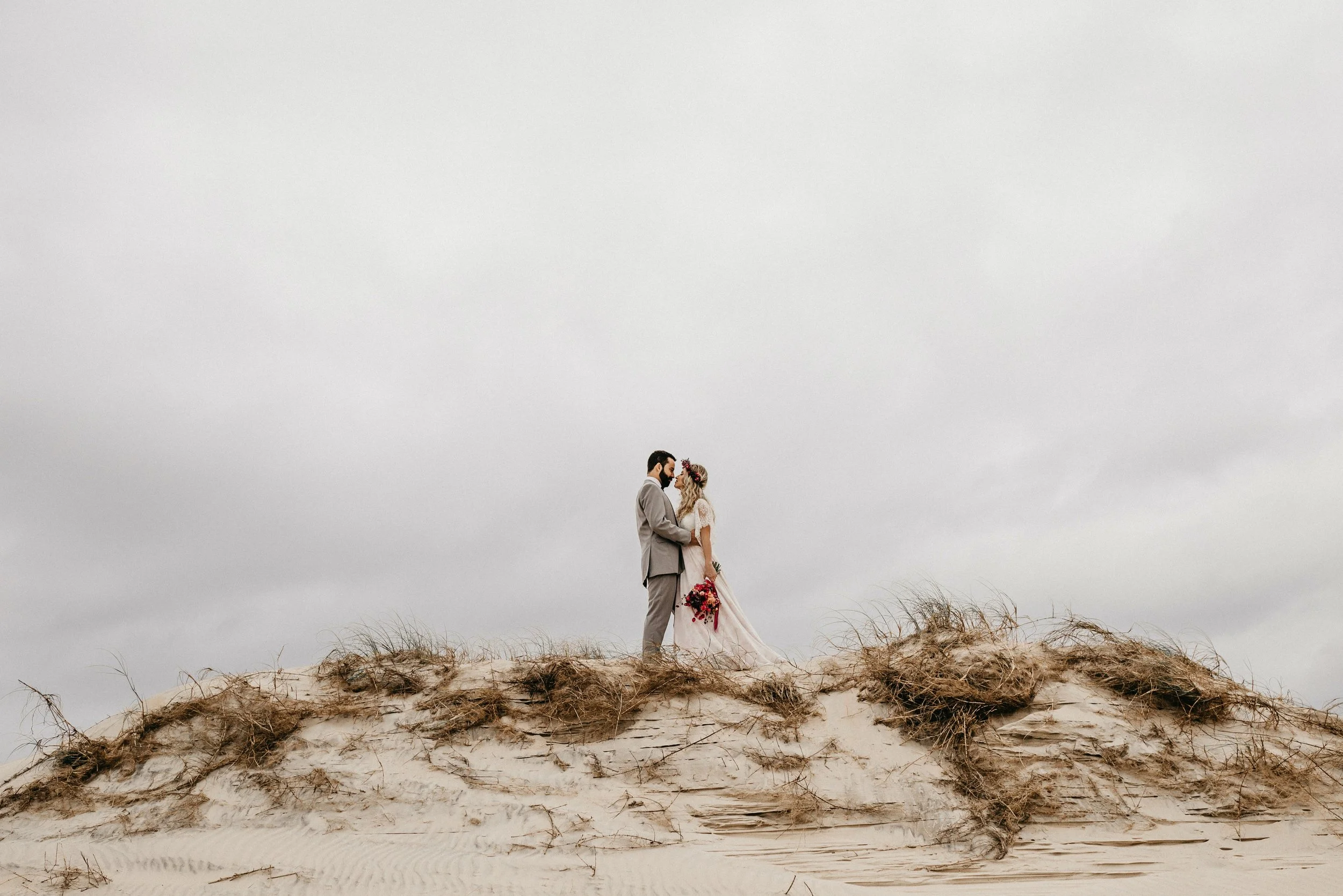 A bride and groom standing close together on a sandy dune landscape under a cloudy sky, holding each other and gazing into each other's eyes.