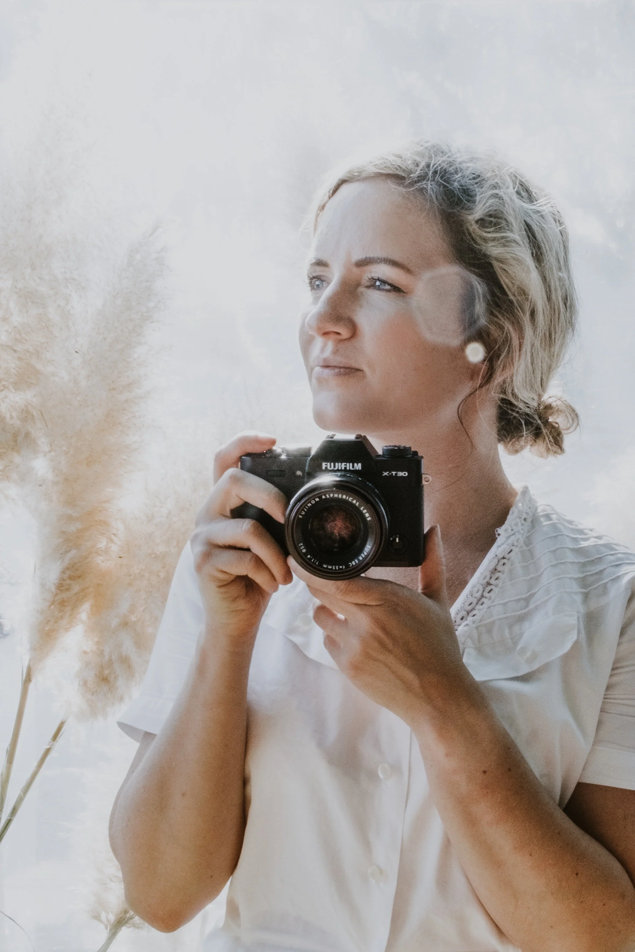 A woman with short blonde hair holding a camera, taking a mirror selfie, wearing a white shirt, standing next to a tall beige pampas grass plant.
