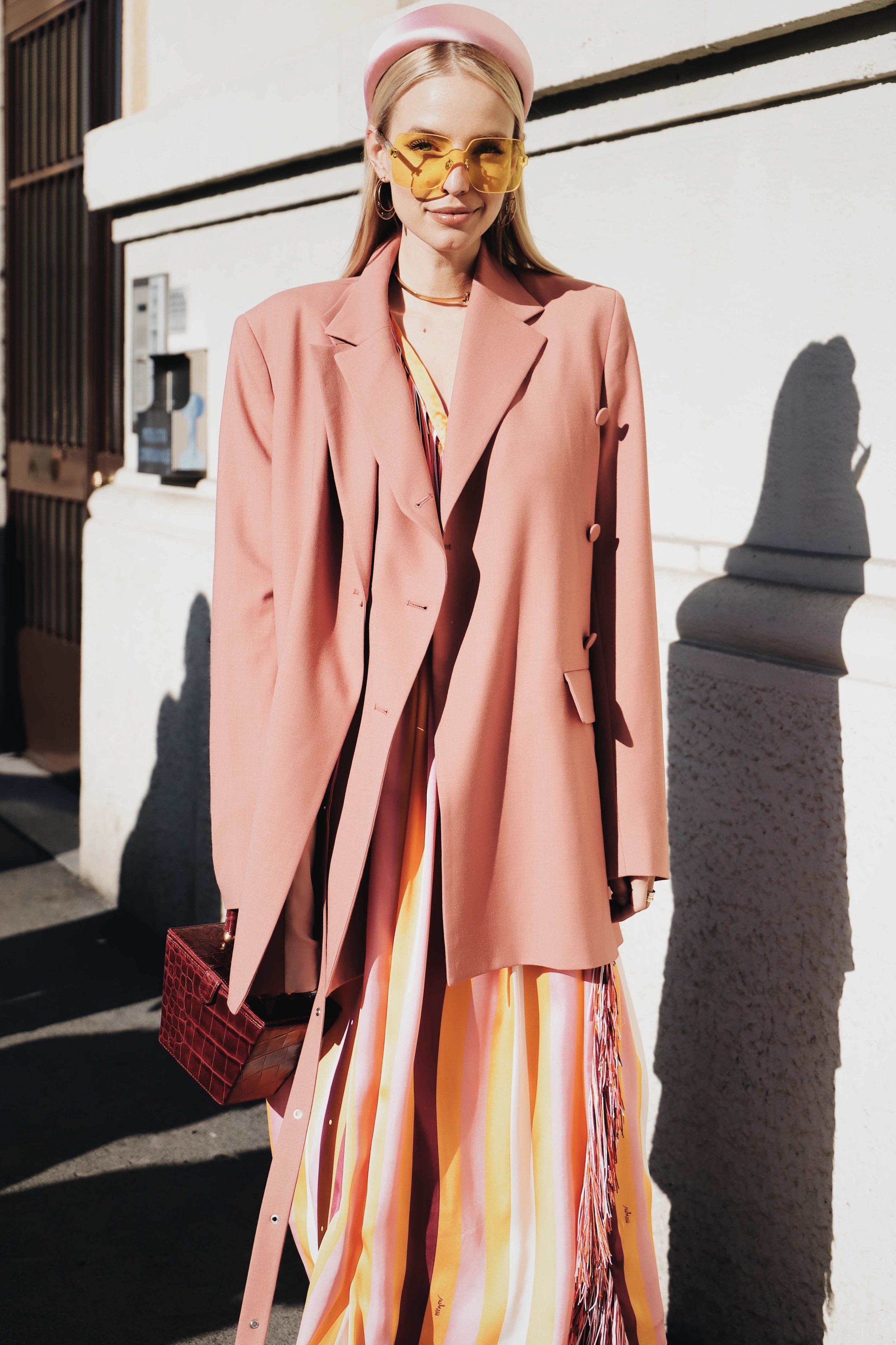 A woman dressed in a pink blazer, a pink hat, yellow-tinted sunglasses, and a colorful striped dress walking on a sunny street, holding a red handbag.