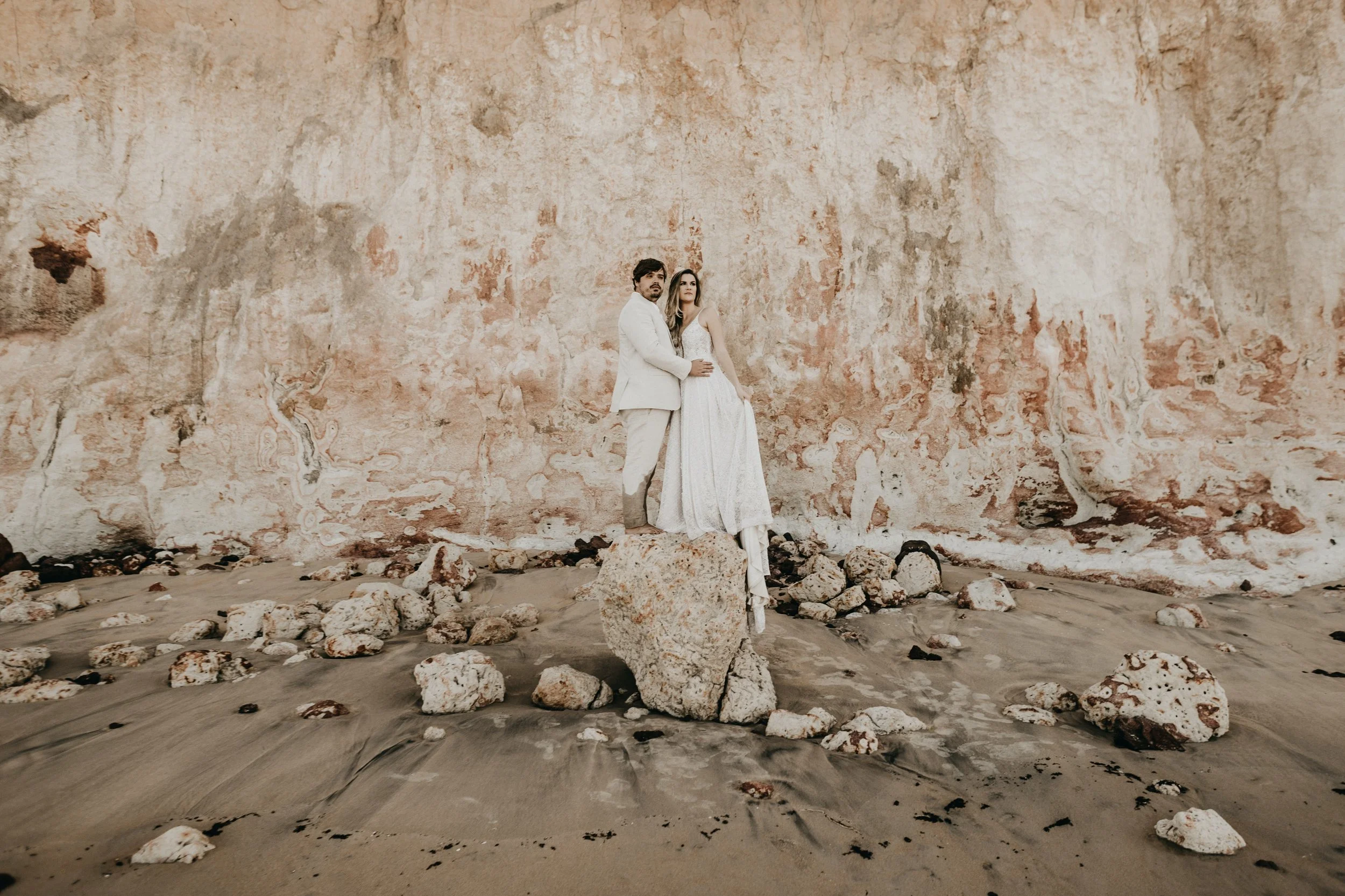 A couple dressed in wedding attire standing in front of a large weathered wall on a rocky beach.