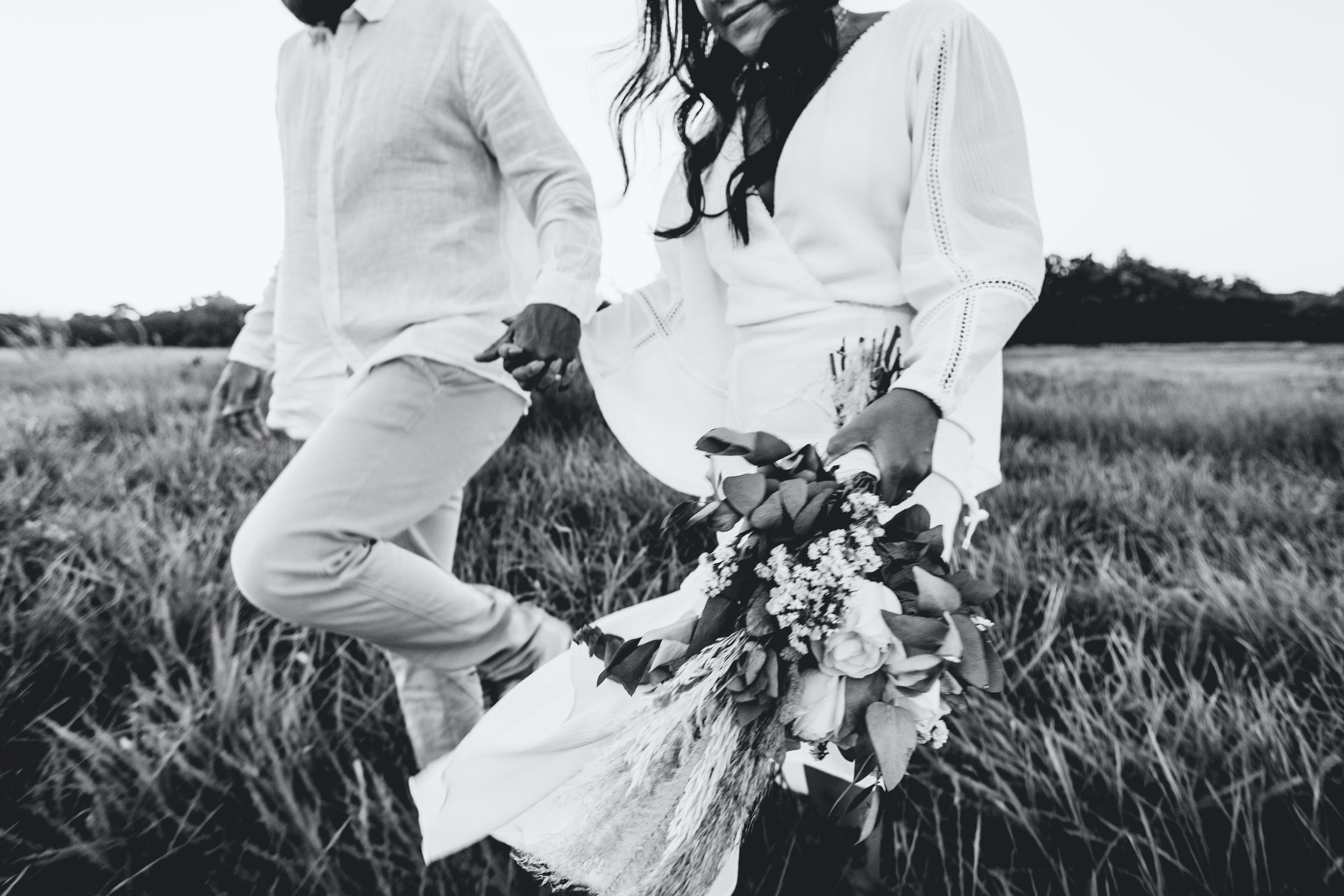 A black and white photo of a man and woman holding hands outdoors. The woman is holding a bouquet of flowers. They are dressed in light-colored clothing, and the background shows an open field with trees in the distance.