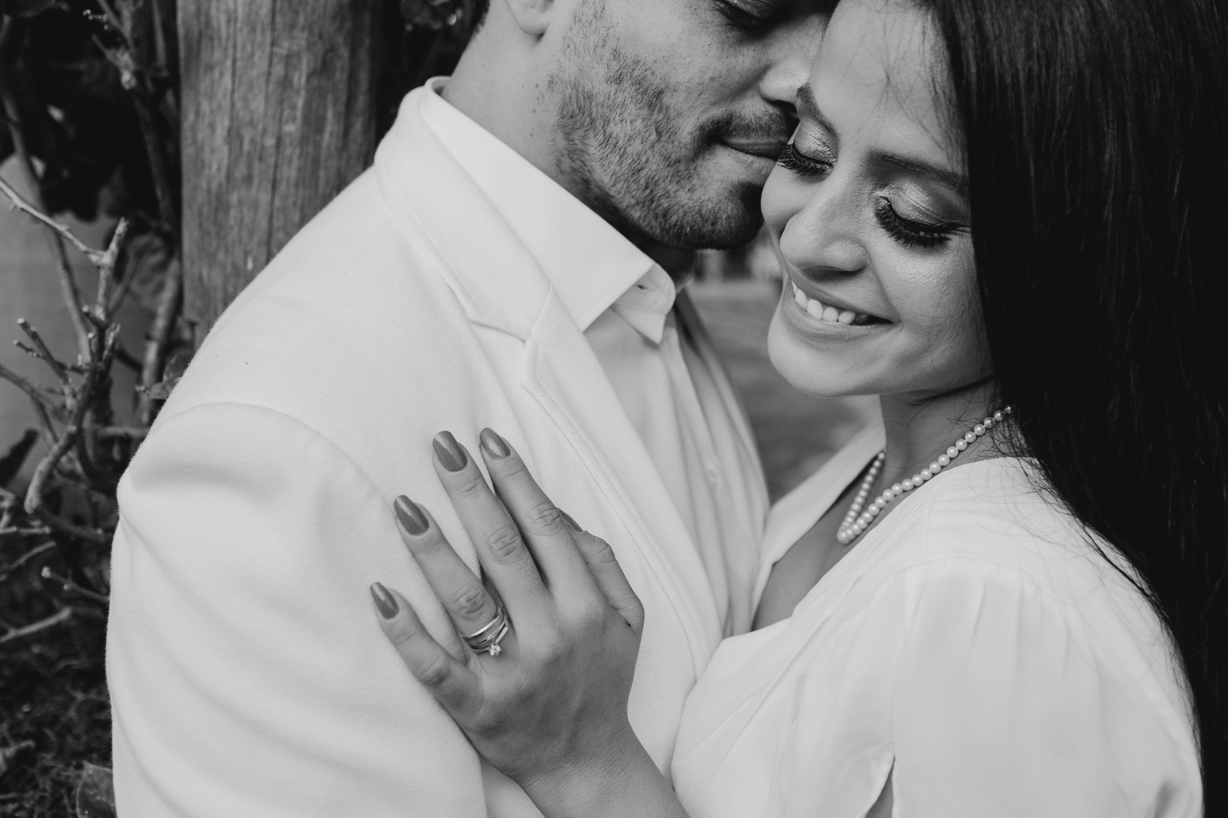 A close-up black-and-white photograph of a couple in an intimate moment, with their faces close together and their eyes closed. The woman has dark hair, wears eyeshadow, and a pearl necklace. The man has short hair and a beard.