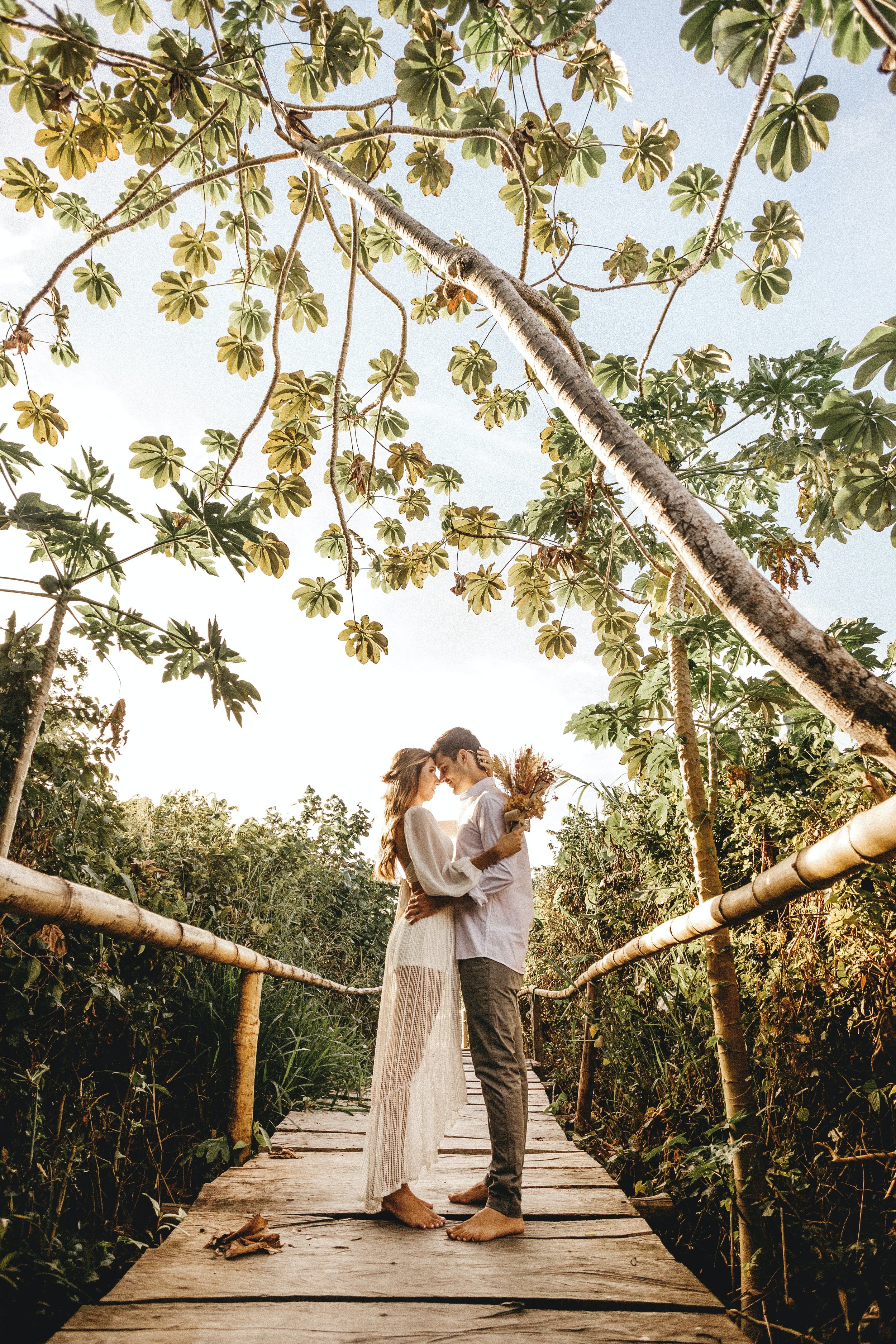A couple standing barefoot on a wooden bridge surrounded by lush green foliage, holding hands and touching foreheads, with sunlight filtering through the trees.