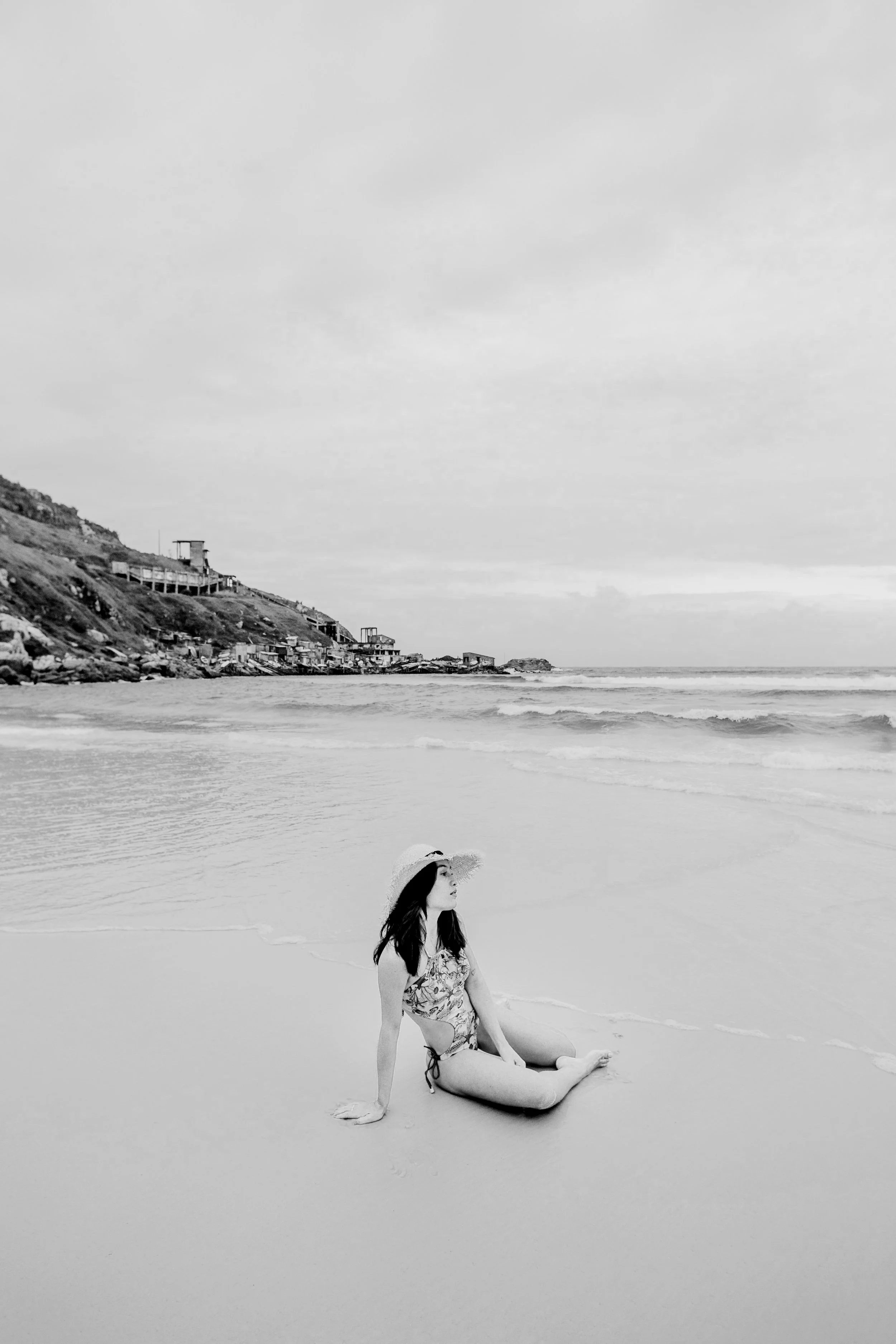 A woman with long dark hair sitting on the wet sand at the beach, wearing a floral swimsuit and a wide-brimmed straw hat, looking to her left with waves approaching the shore. In the background, there are buildings on a hillside and cloudy skies.