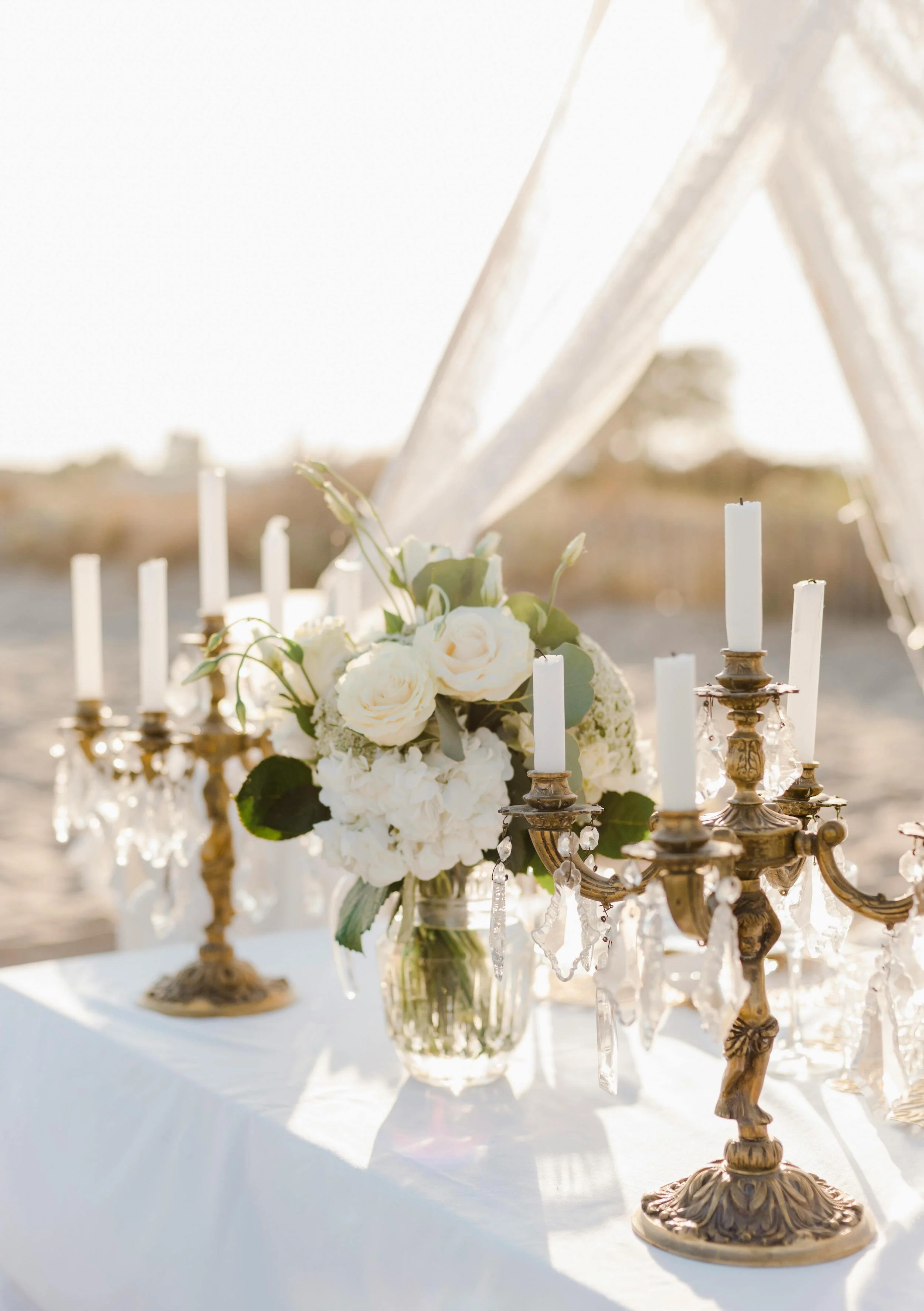 A wedding centerpiece with white roses, hydrangeas, and greenery in a glass vase, surrounded by white candles in ornate gold candelabras decorated with crystal prisms, set outdoors in natural light.