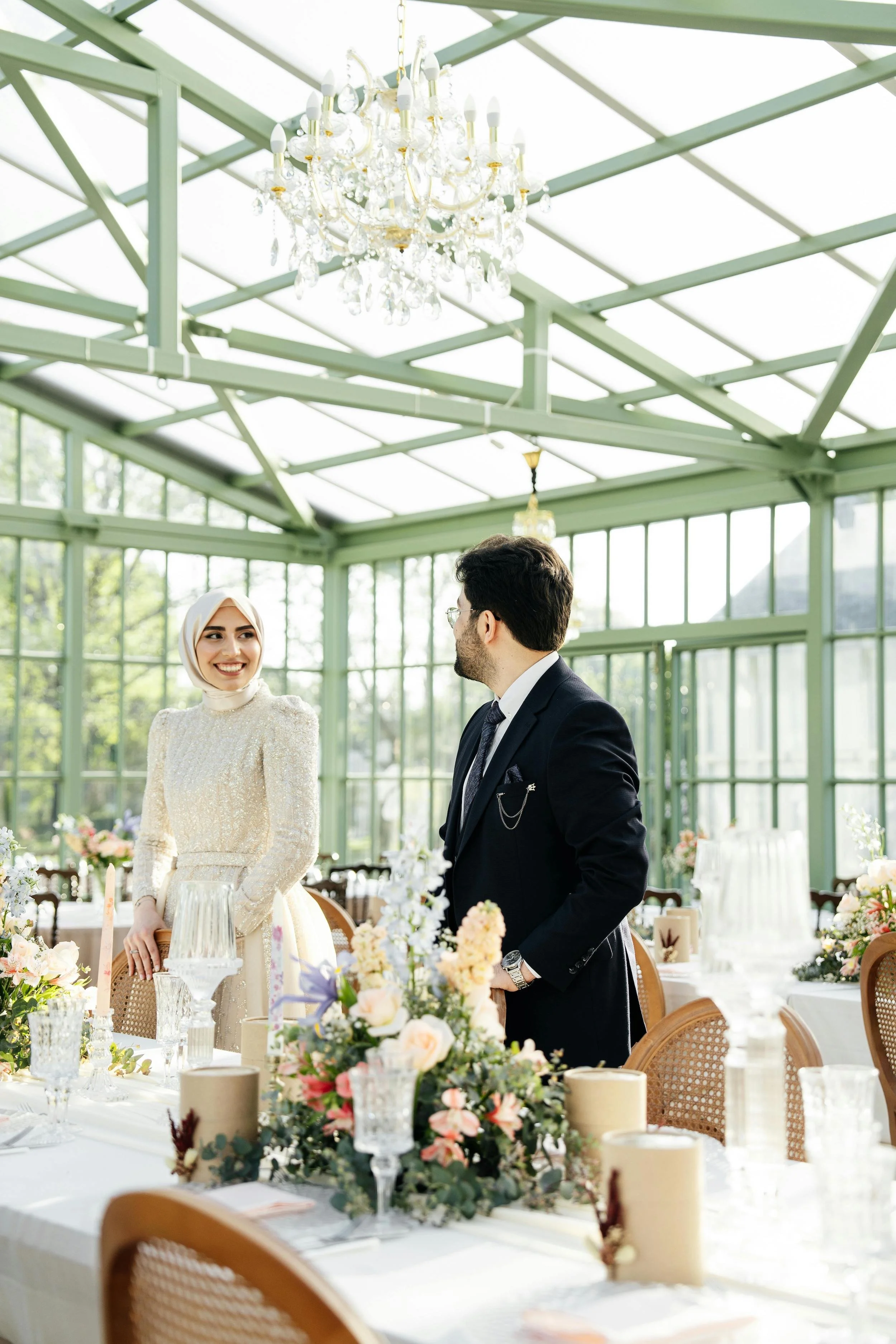 A smiling woman in a wedding dress and hijab stands next to a man in a tuxedo at a wedding reception inside a glass greenhouse, with a decorated table in the foreground and chandelier hanging from the ceiling.