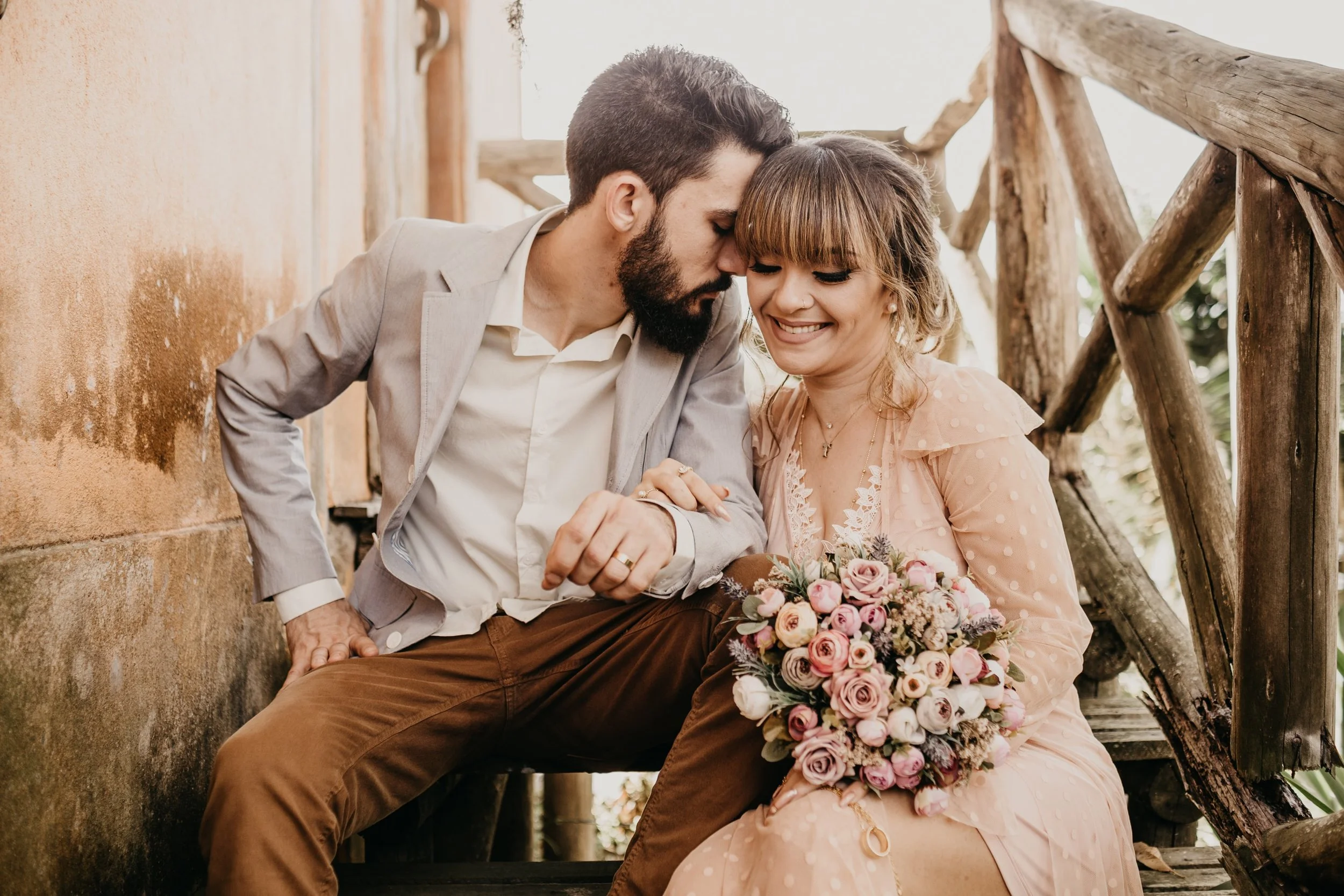 A couple sitting close together on a wooden bench, with the man leaning his forehead against the woman's. The woman is holding a bouquet of pink and white roses and smiling. They are outdoors near a rustic wooden railing and an orange wall.