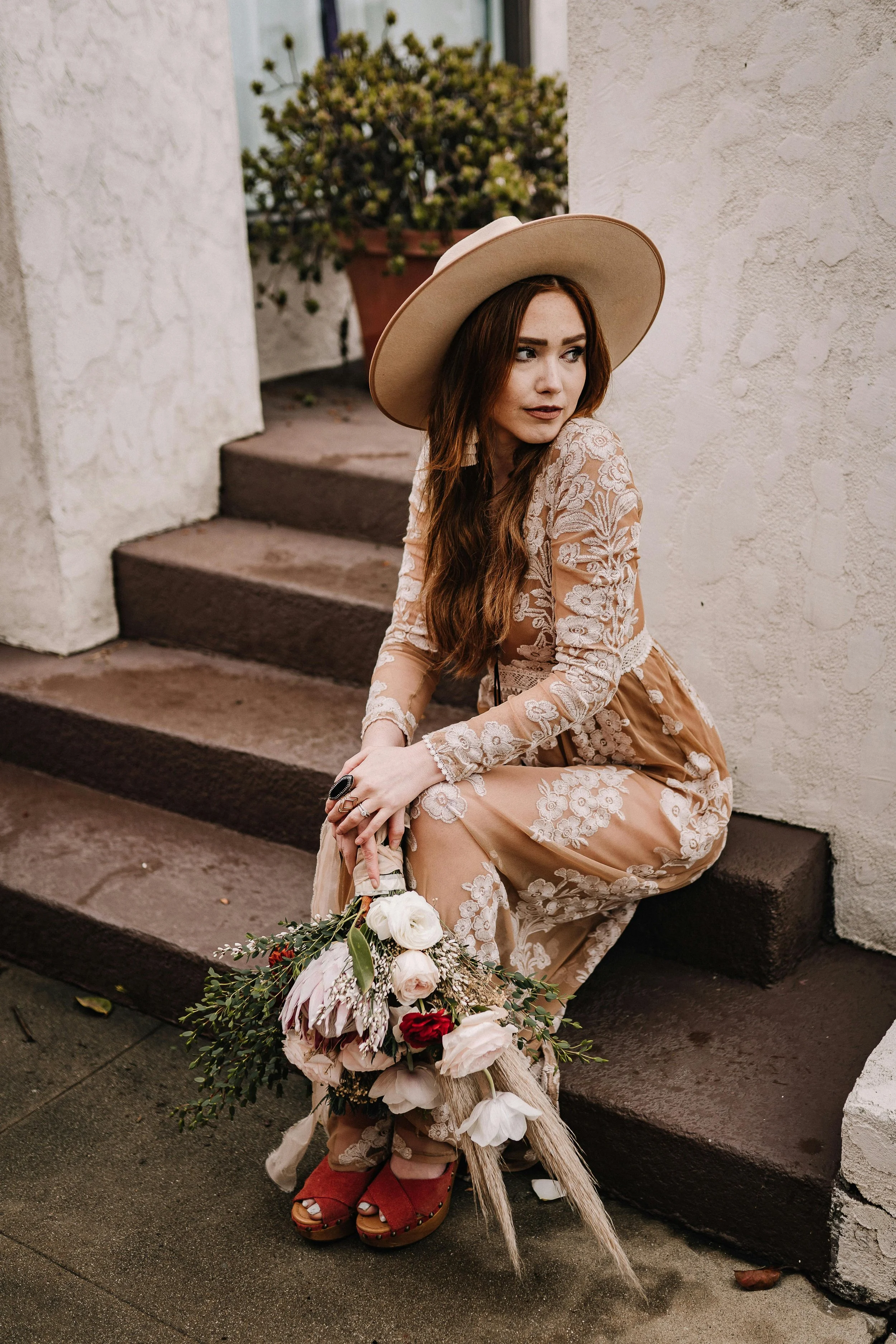 A young woman with long brown hair, fair skin, and dark lipstick, wearing a beige floral lace dress and a large beige floppy hat. She is sitting on steps outside, holding a bouquet of white and red flowers with greenery. She is looking to her left, with a pensive expression. The background includes white stucco walls, steps, and a large potted plant with light-colored flowers.