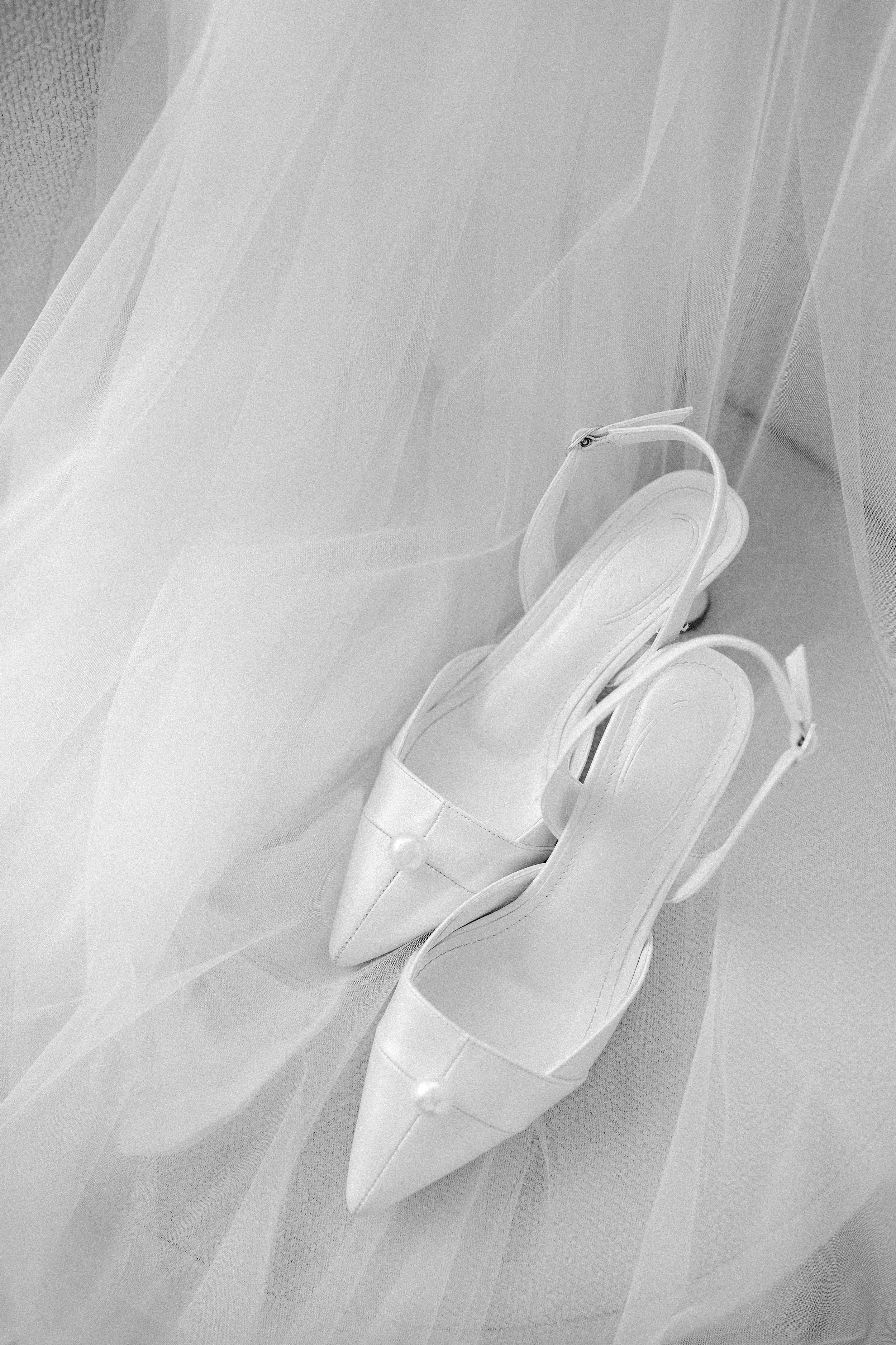 Set of white high-heeled shoes with platform soles and ankle straps, placed on a floor with a sheer fabric curtain in the background.