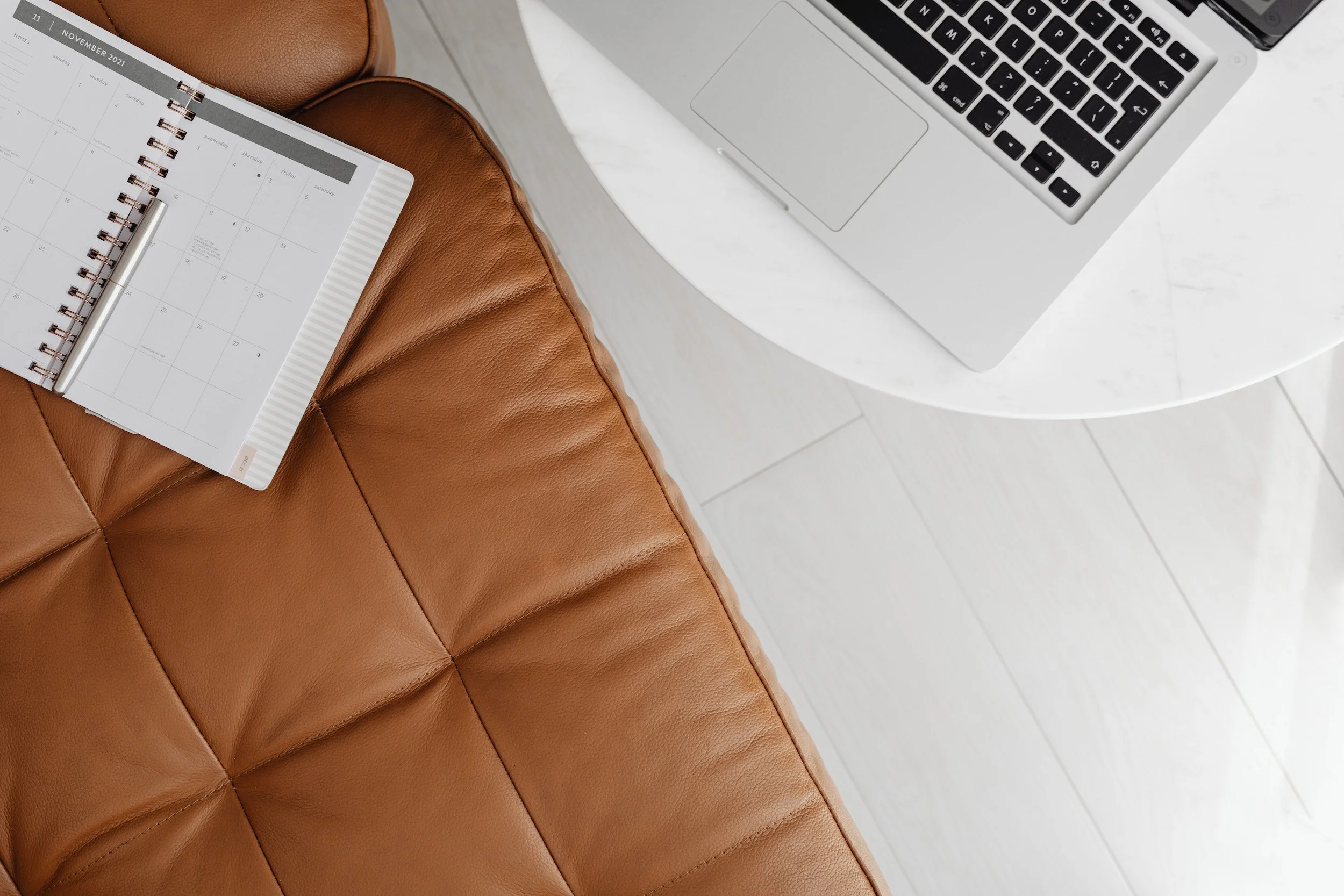 A workspace with a brown leather chair, a white marble table, a silver MacBook laptop, and an open planner for November 2021 with a silver pen resting on it.