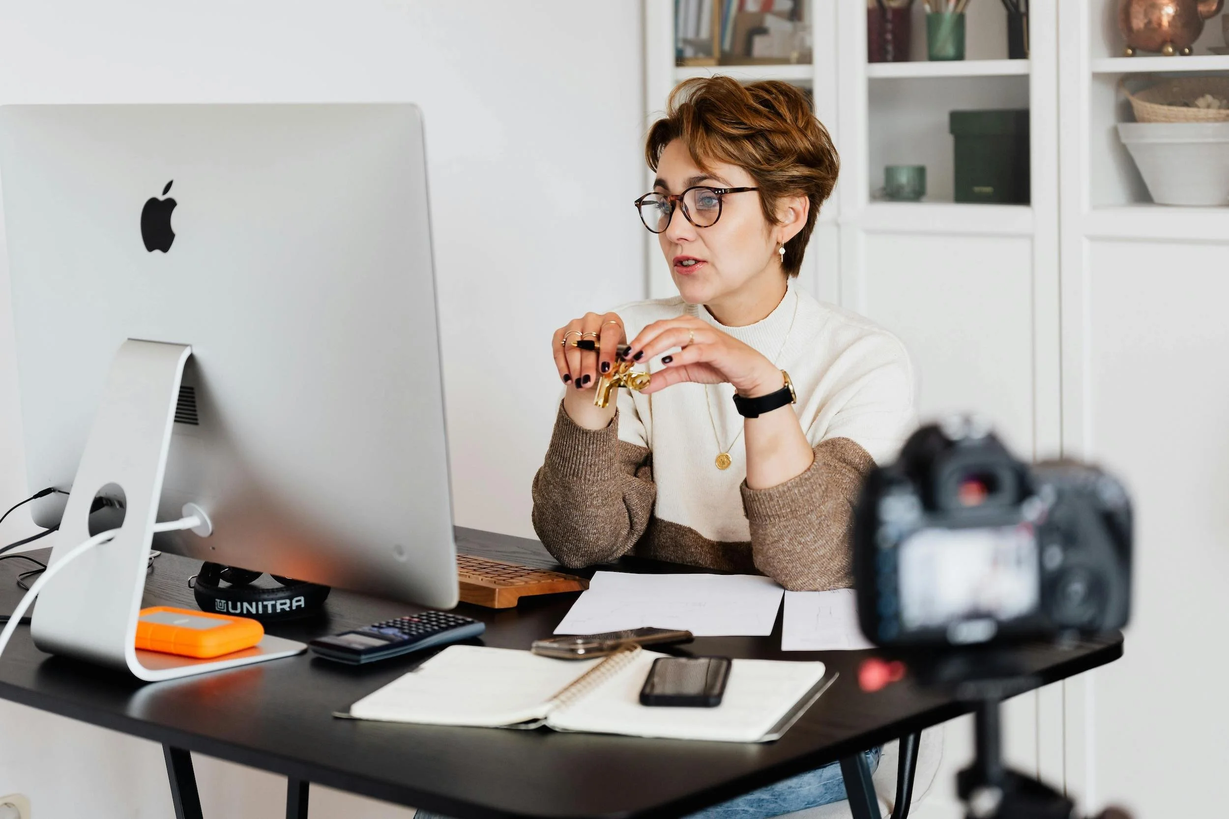 A woman with short brown hair and glasses sitting at a desk with an Apple computer, a notebook, and a camera in a bright room with white shelves.