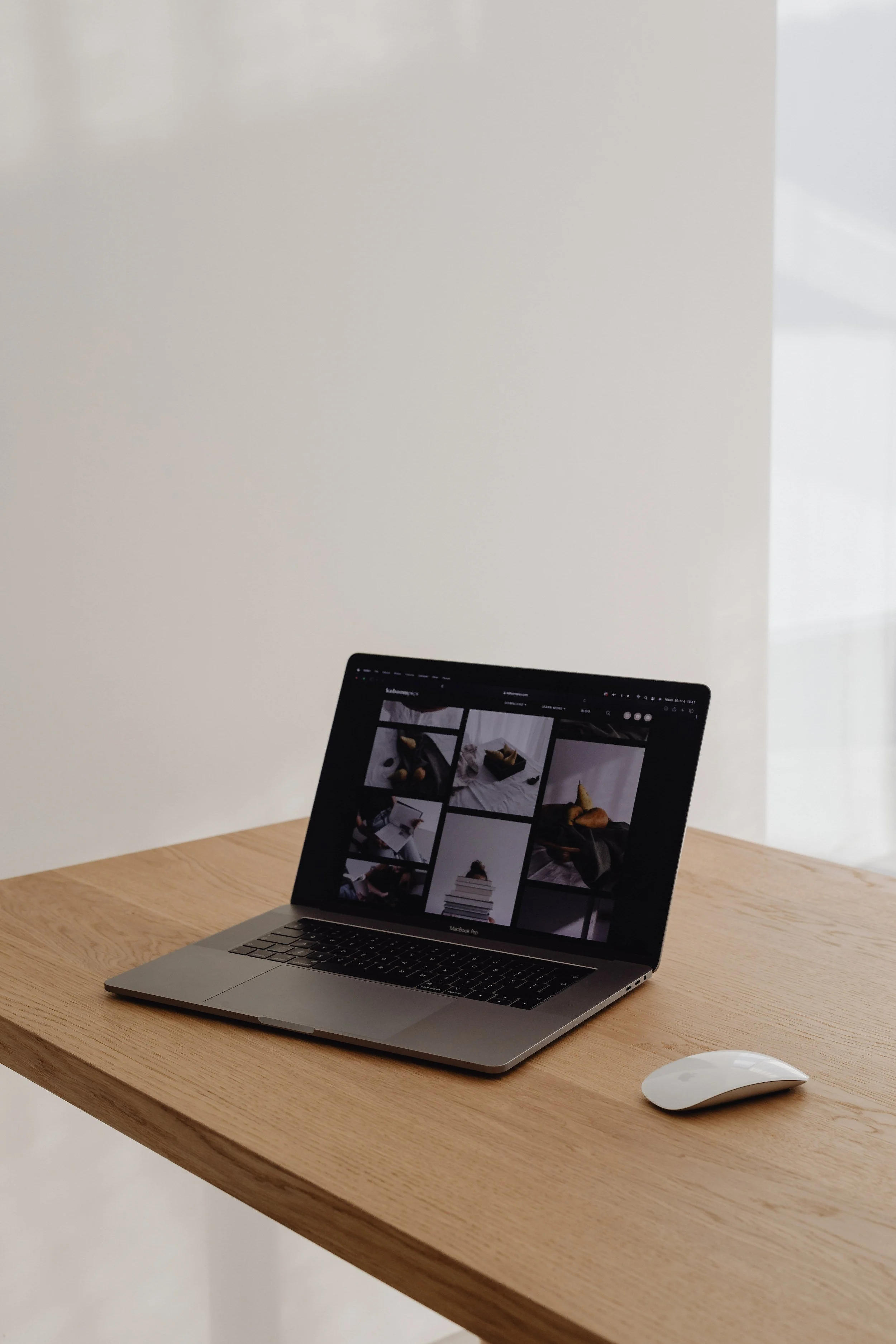 A laptop on a wooden desk displays a grid of images, with a white mouse beside it, against a plain white wall.
