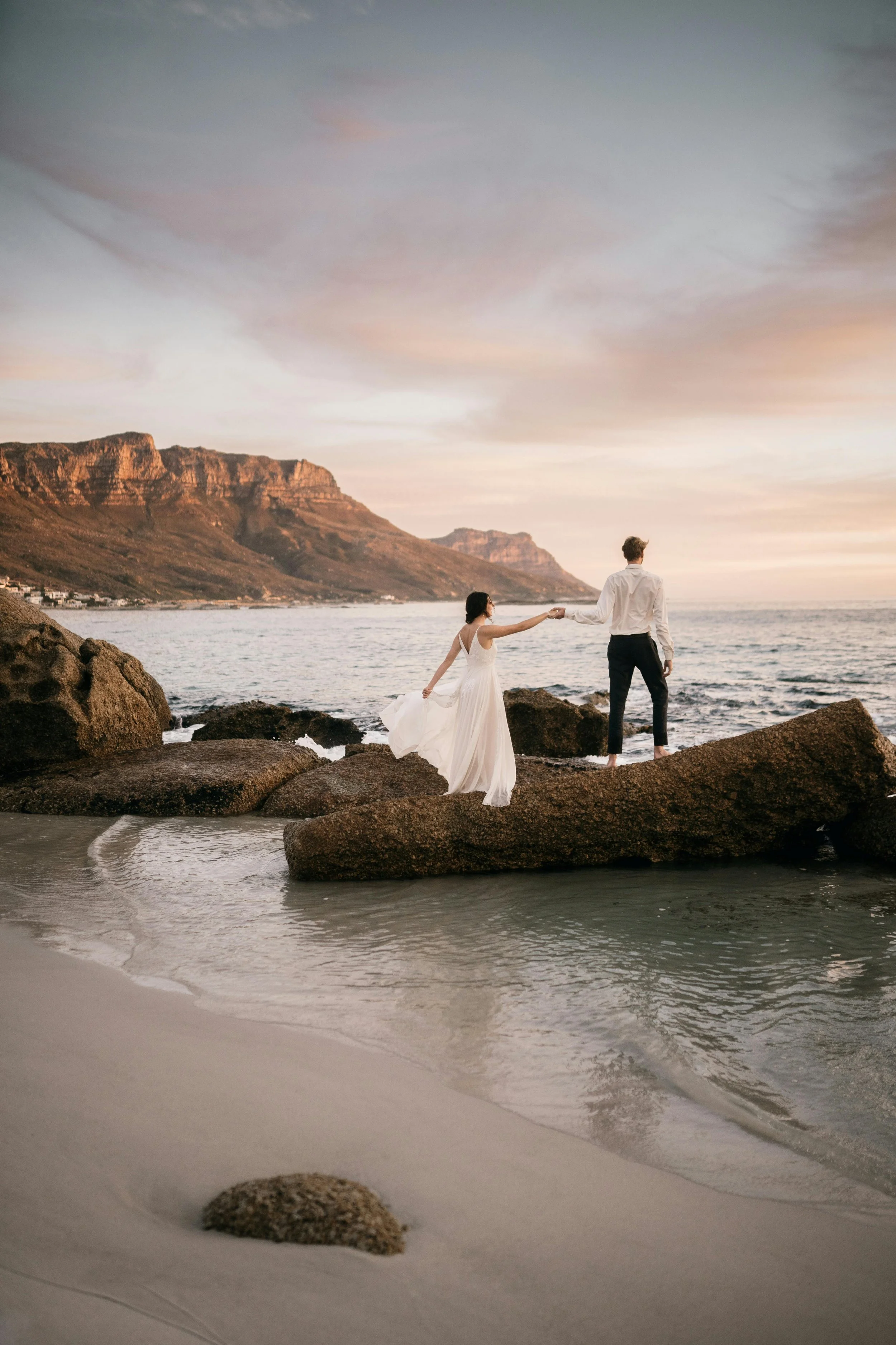 A couple in wedding attire stands on rocks at a beach during sunset, holding hands with water and mountains in the background.