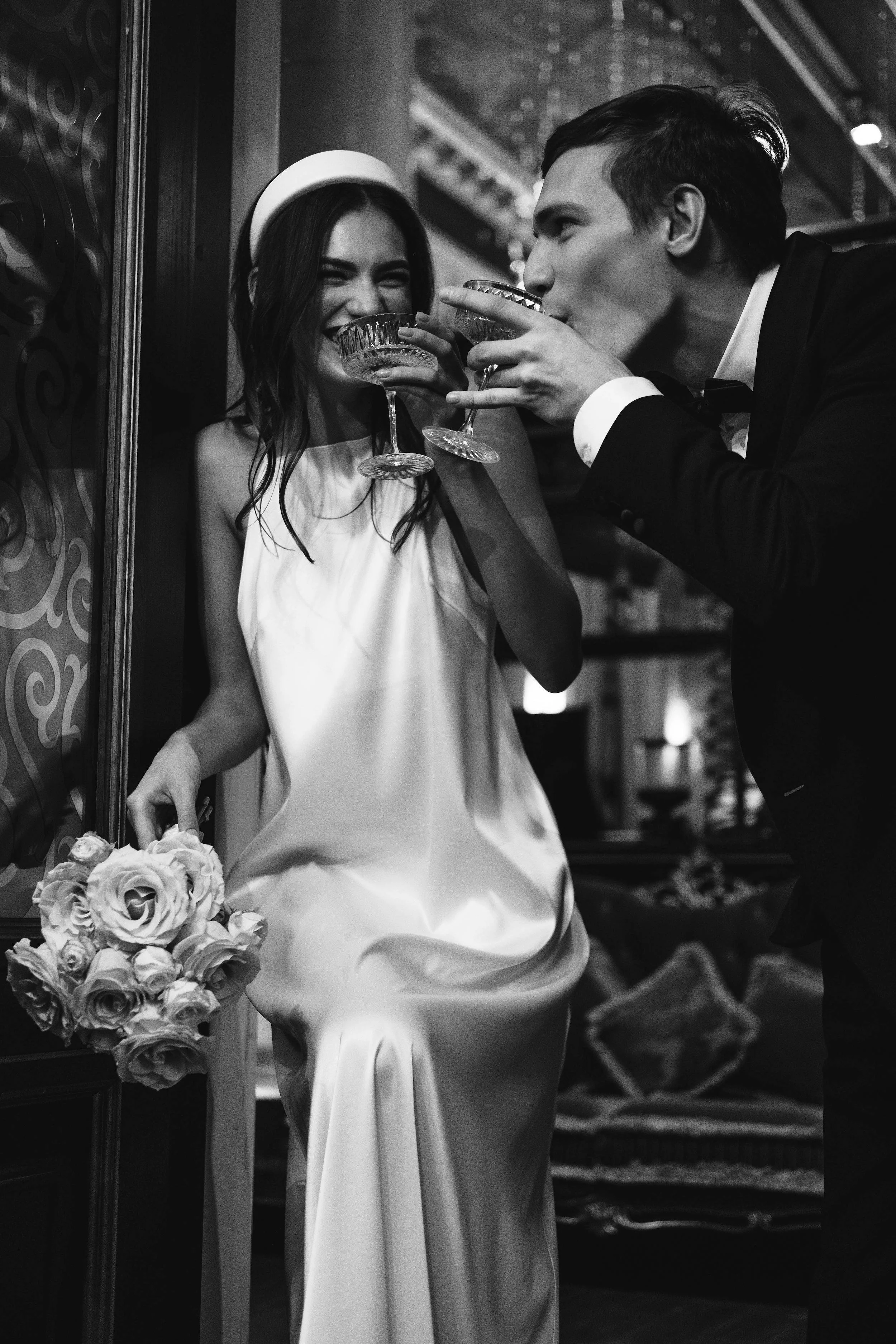 A young woman in a white dress and headband, smiling with eyes closed, holding a bouquet of roses. A man in a tuxedo leans in with a drink, sharing a moment with her.