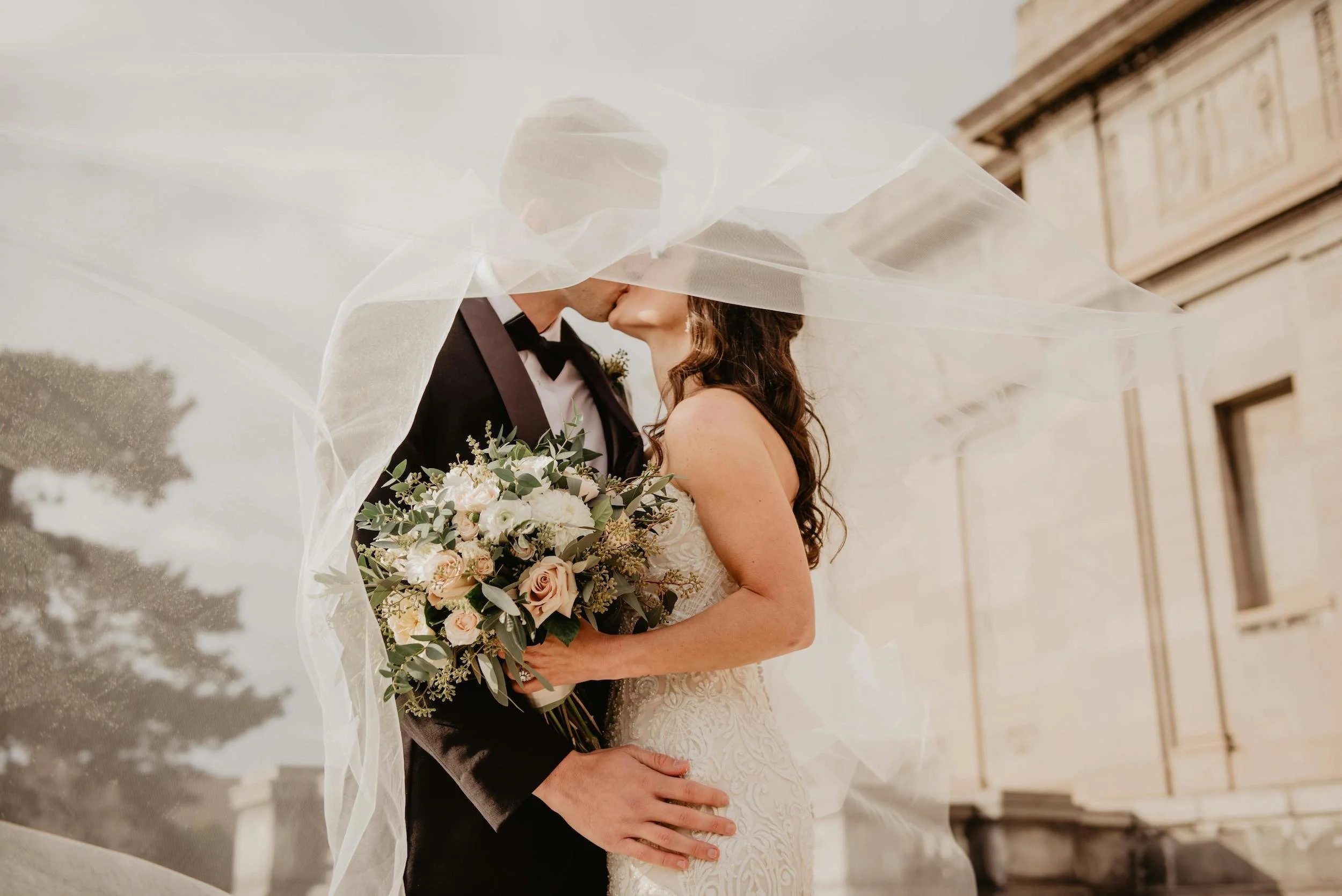 A newlywed couple sharing a kiss, with the bride holding a bouquet of flowers, under a flowing white bridal veil on a city street near historic buildings.