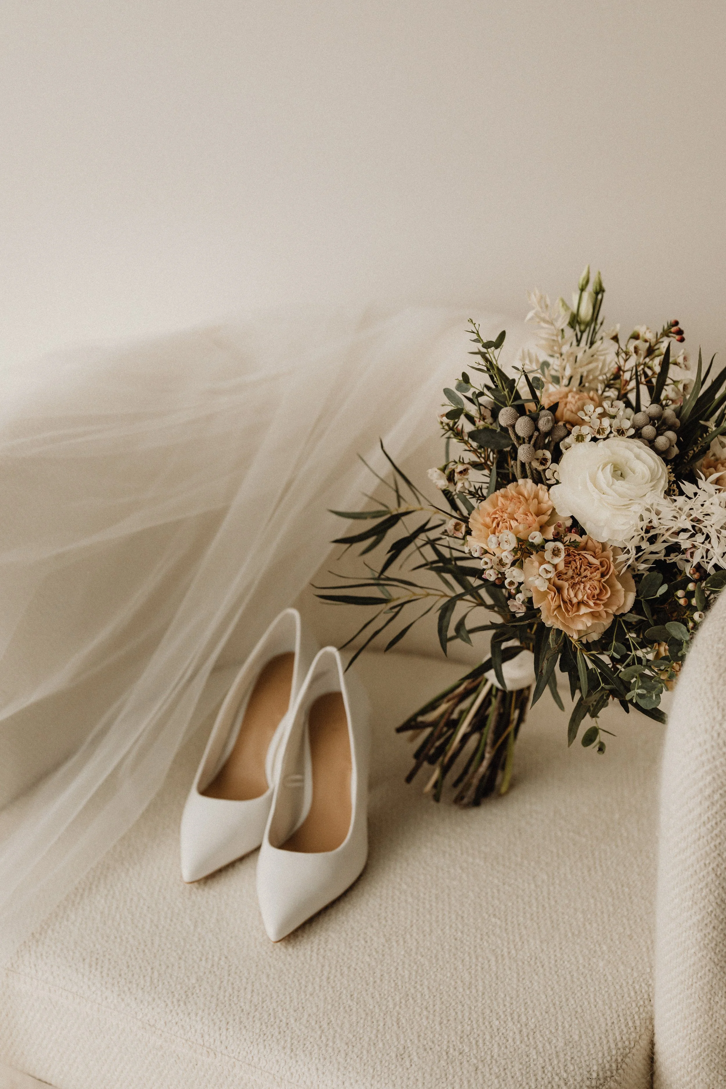 A bouquet of mixed flowers including white roses, peach carnations, and greenery, placed next to a pair of white high heels on a beige textured surface.