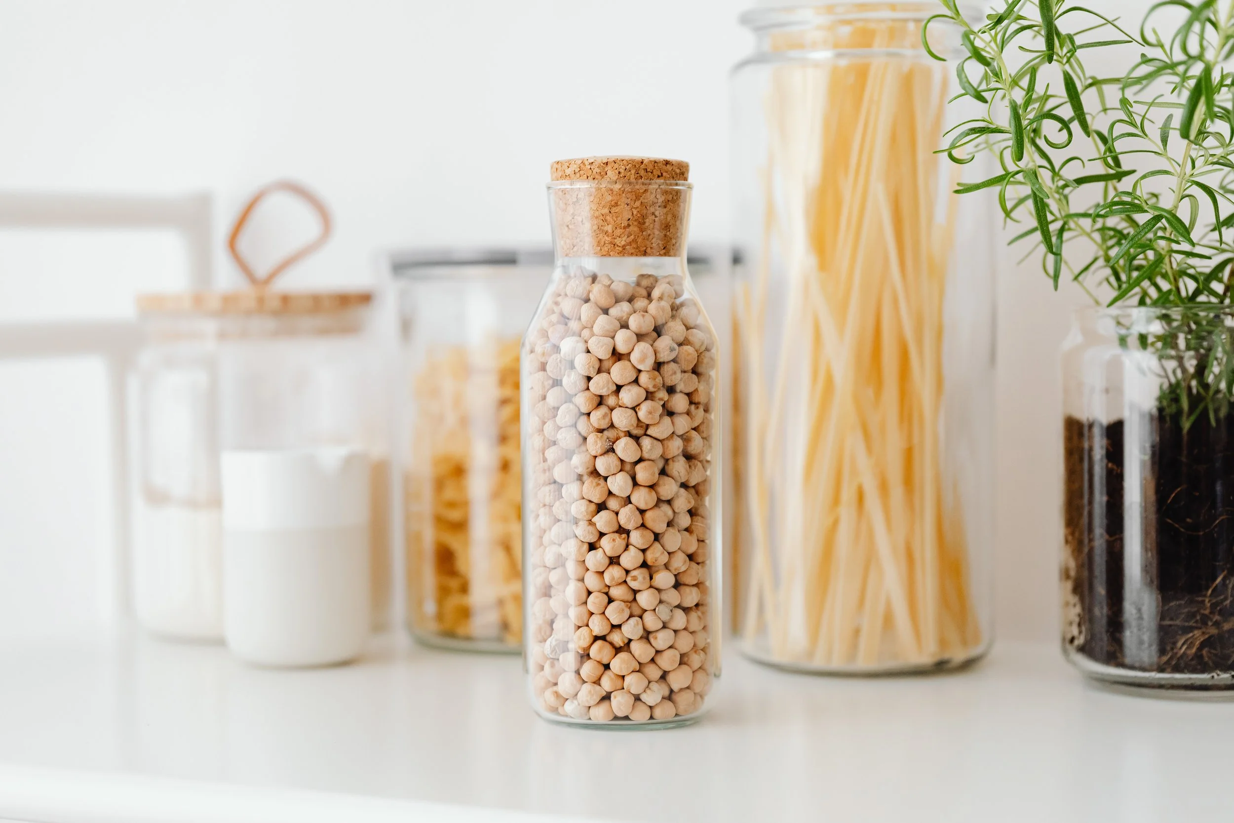 Clear glass jars filled with pasta, chickpeas, and spices, with green herbs in a container on the right, arranged on a white surface.