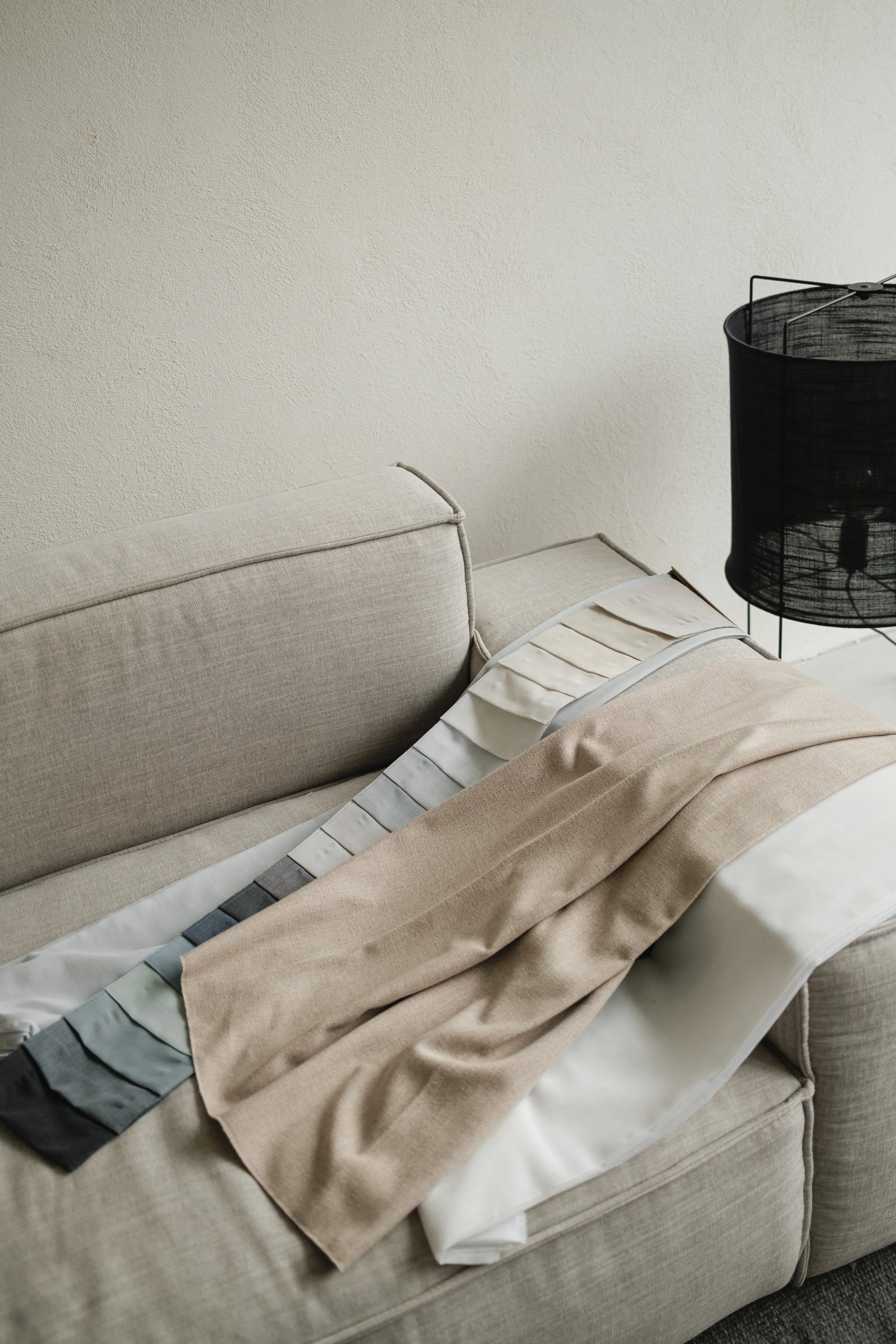 A beige couch with a stack of fabric samples or swatches spread across the armrest, next to a black lamp with a cylindrical shade, against a plain off-white wall.