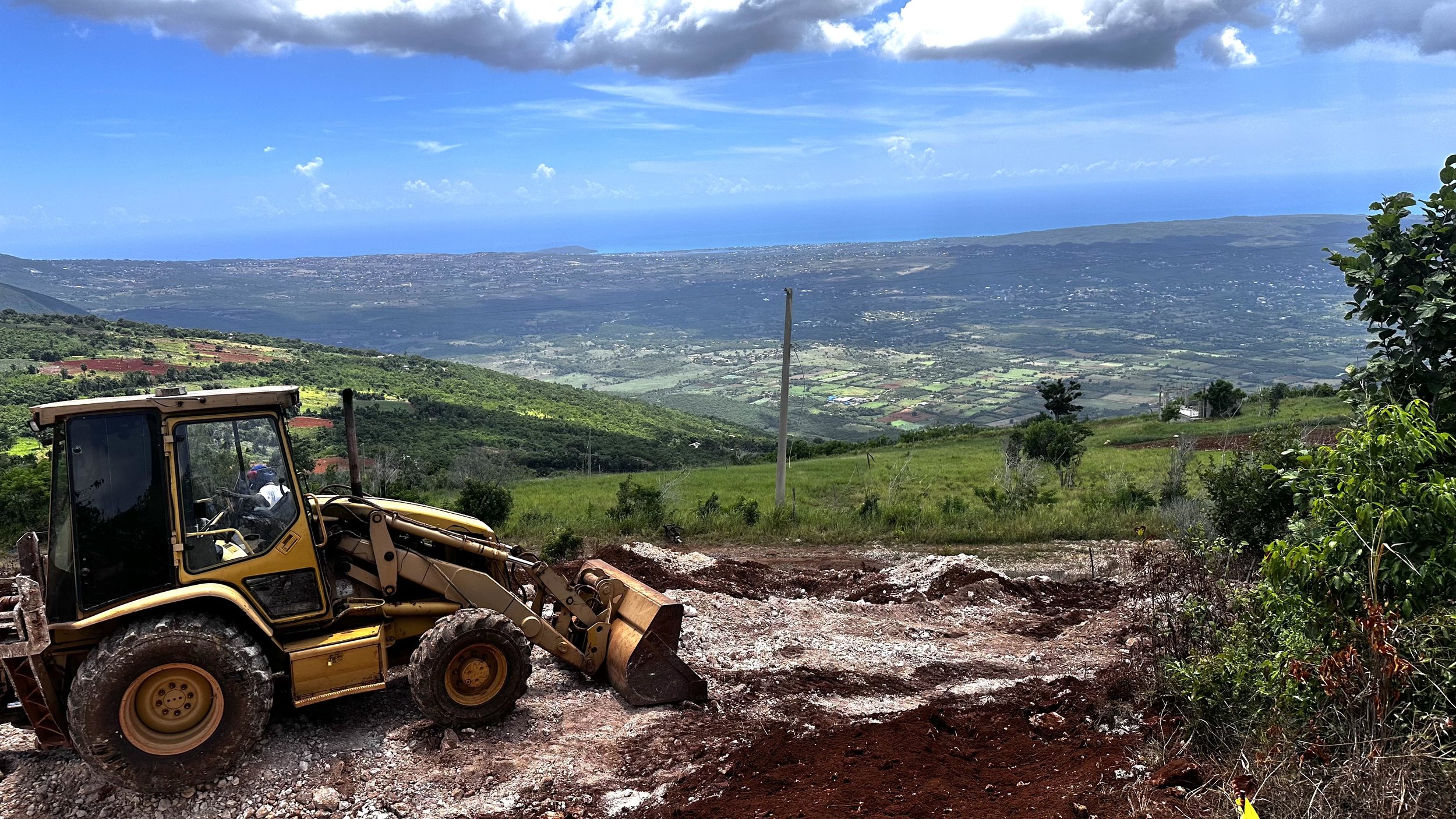 A yellow bulldozer working on a hillside foreground with a scenic view of the valley, ocean, and sky in the background.